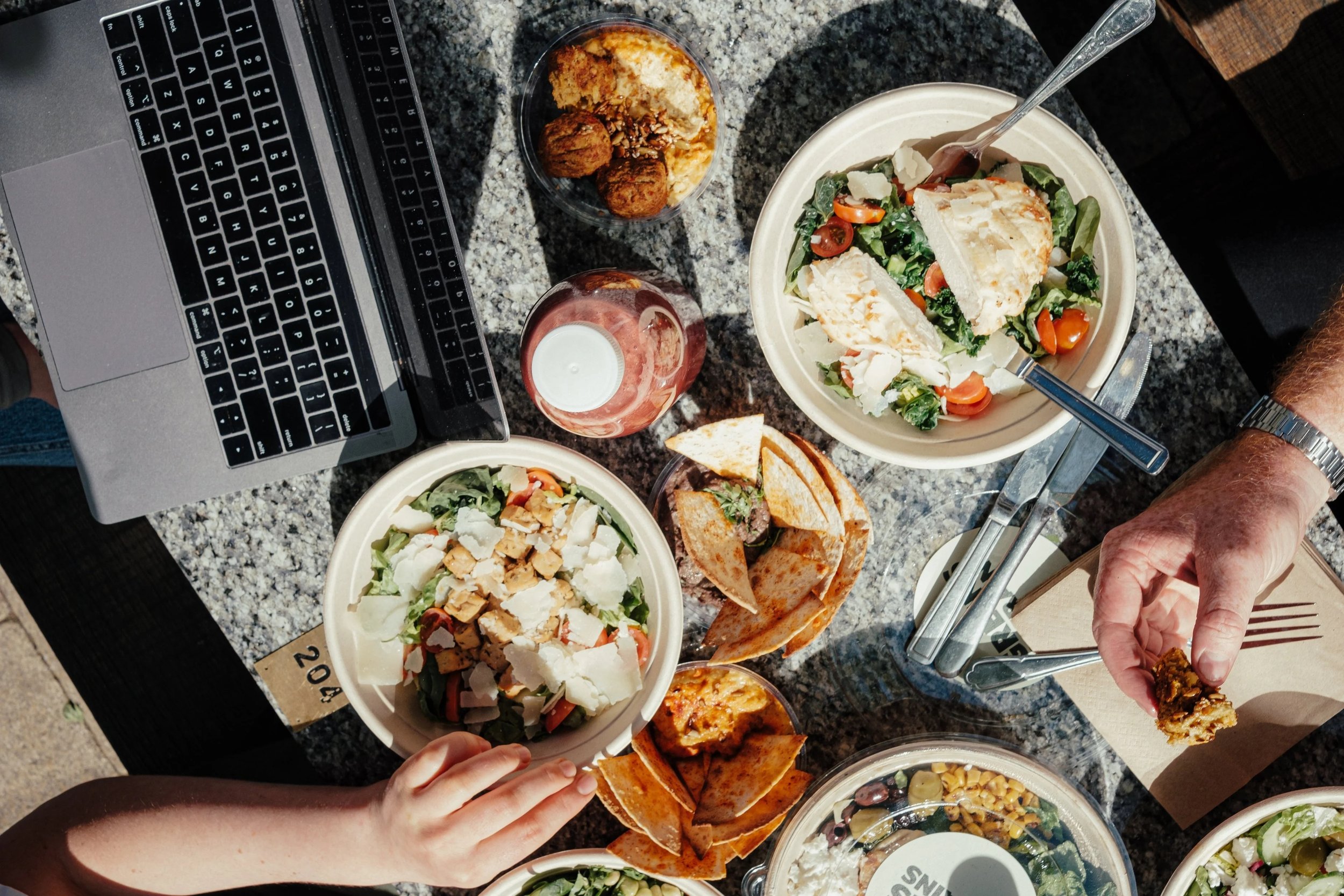 Greens & Grains being shared. A table with multiple dishes including salads with shredded chicken, cherry tomatoes, and cheese, grilled sandwiches, hummus with pita chips, and two drinks, with a laptop and a person's hand holding a fork visible.