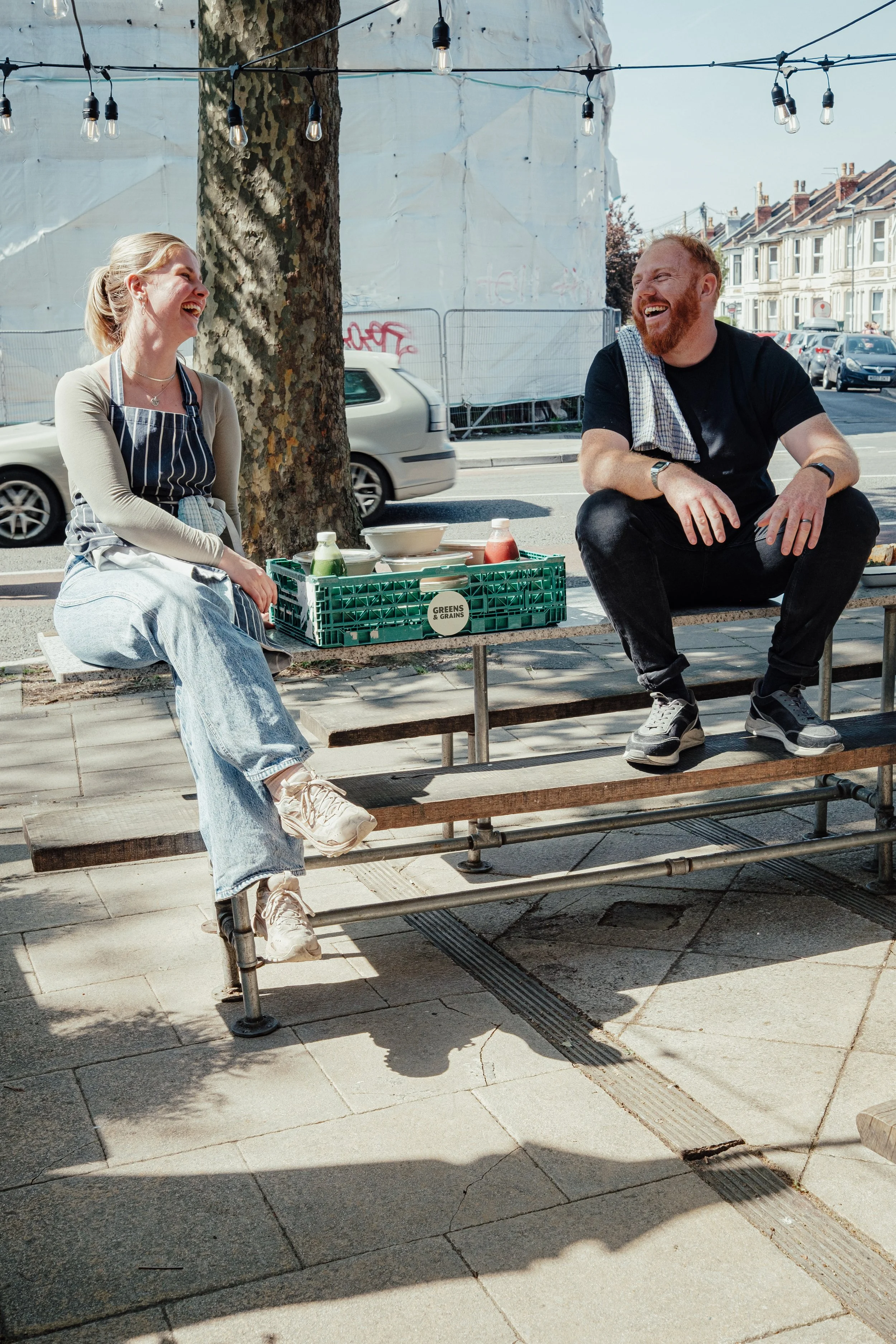 Jack & Mads Co-Founders of Greens & Grains. Two people sitting on a wooden bench outdoors, laughing and talking, with a table holding condiments and bowls in front of them, under string lights near a tree on a city street.
