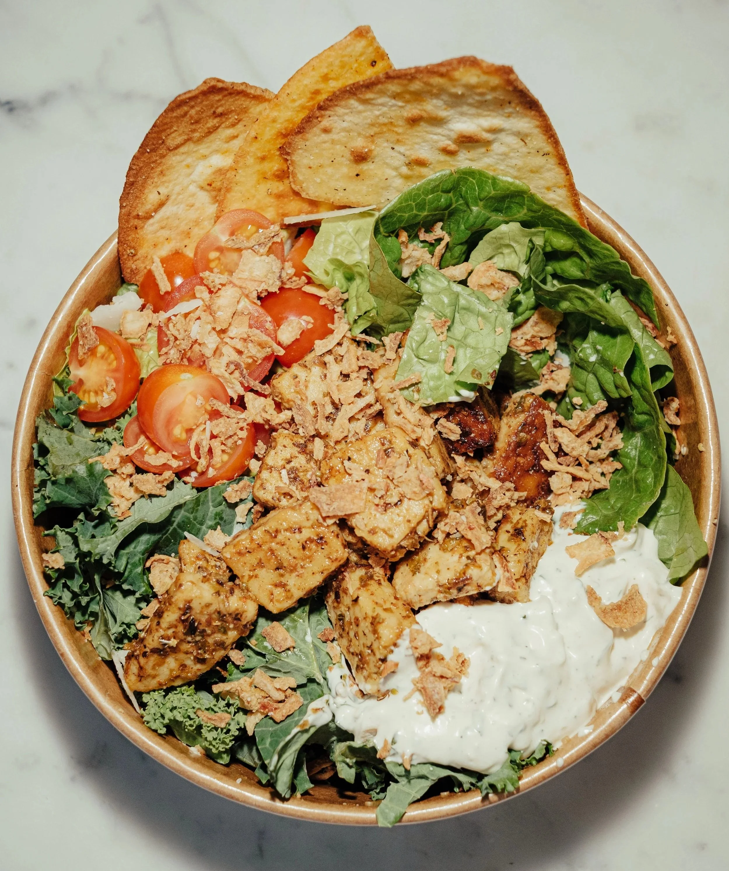 Greens & Grains salad bowl. A bowl of salad with cherry tomatoes, shredded chicken, crispy tortilla chips, leafy greens, and a creamy dressing, topped with crushed tortilla chips.