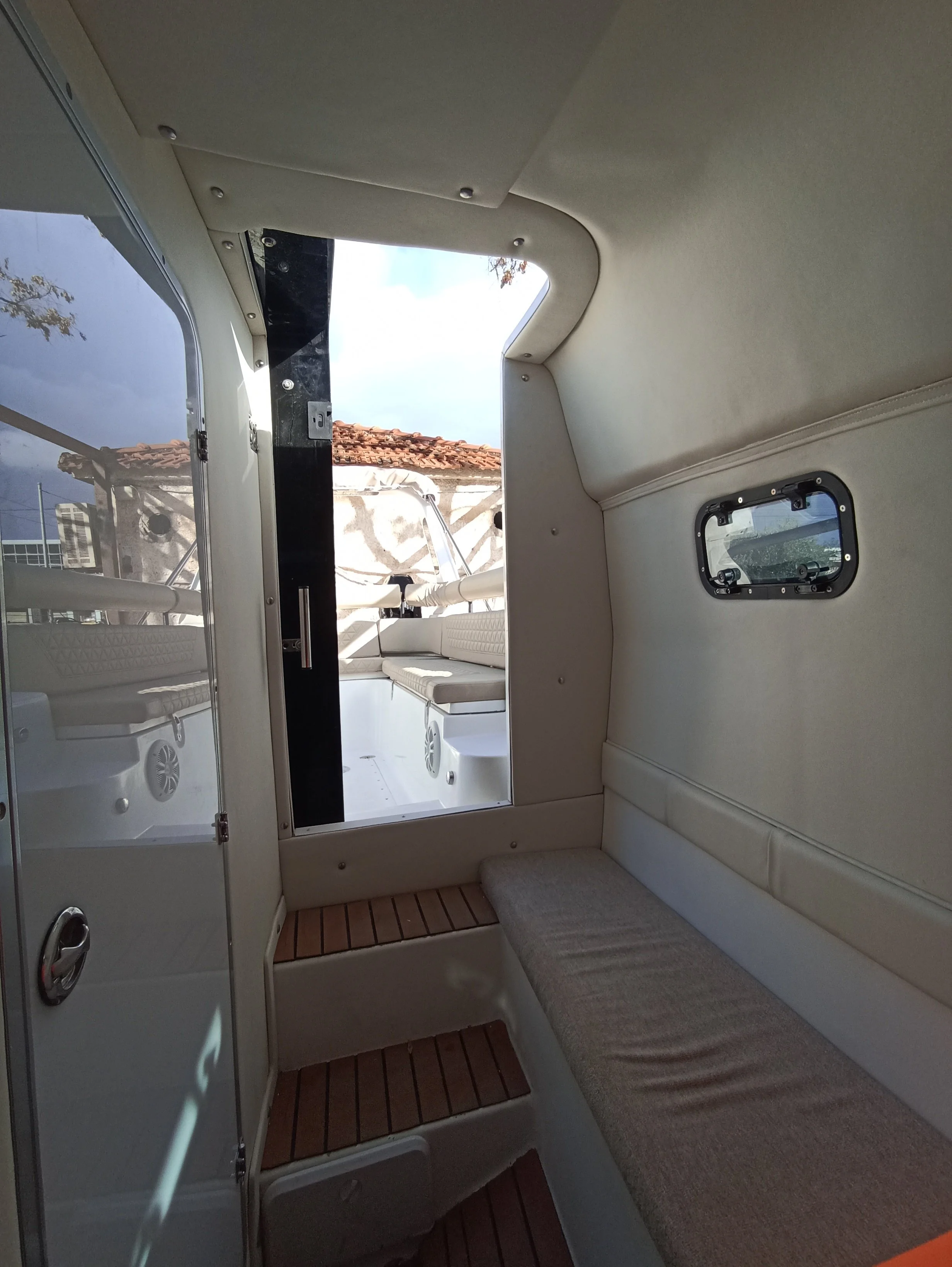 View inside a boat cabin showing a cushioned bench, small windows, and an opening to the outside deck where another boat and some buildings are visible.