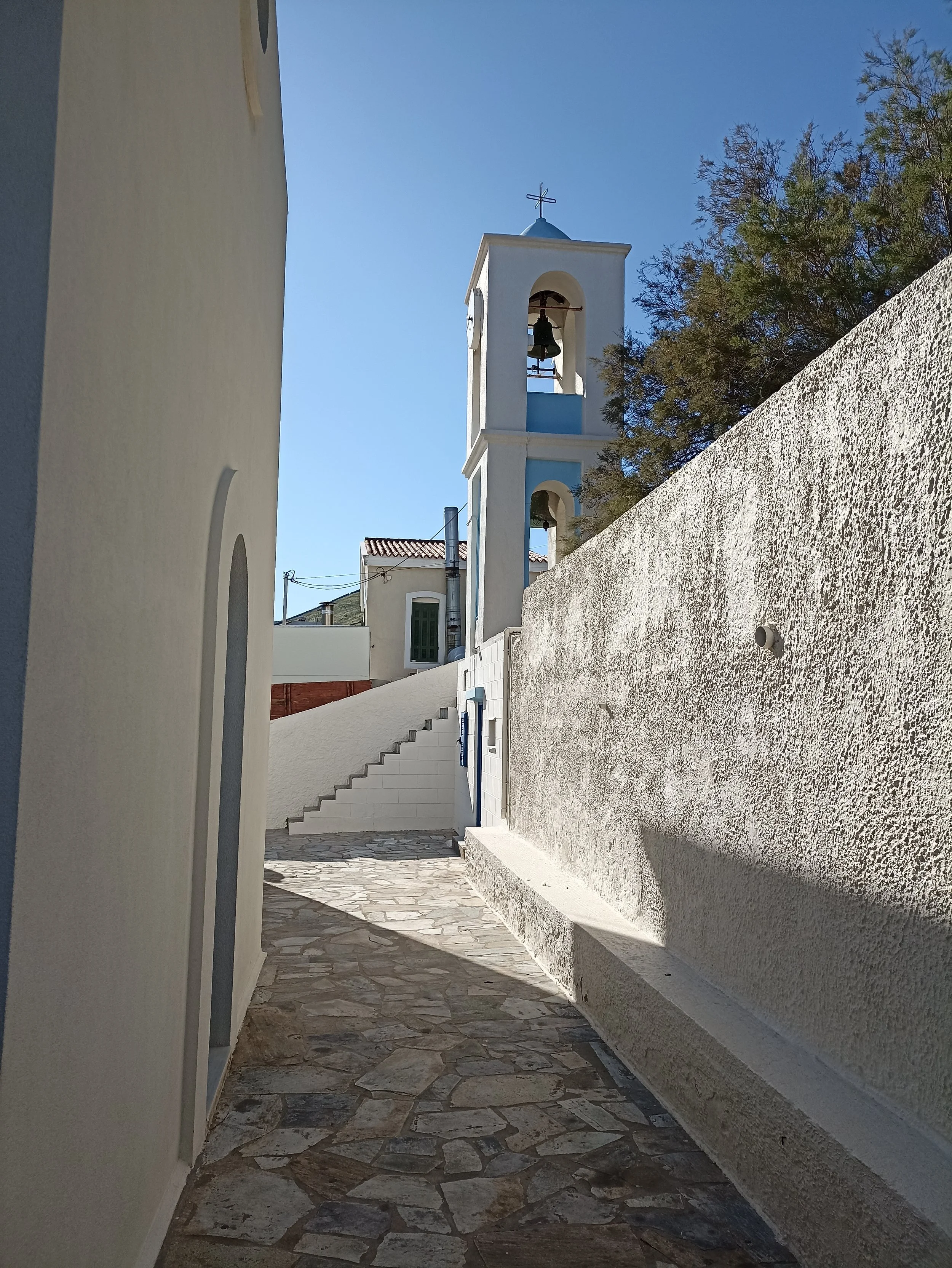 A narrow alleyway with stone paving, white walls, a staircase, and a blue and white church with a bell tower and cross, under a clear blue sky.