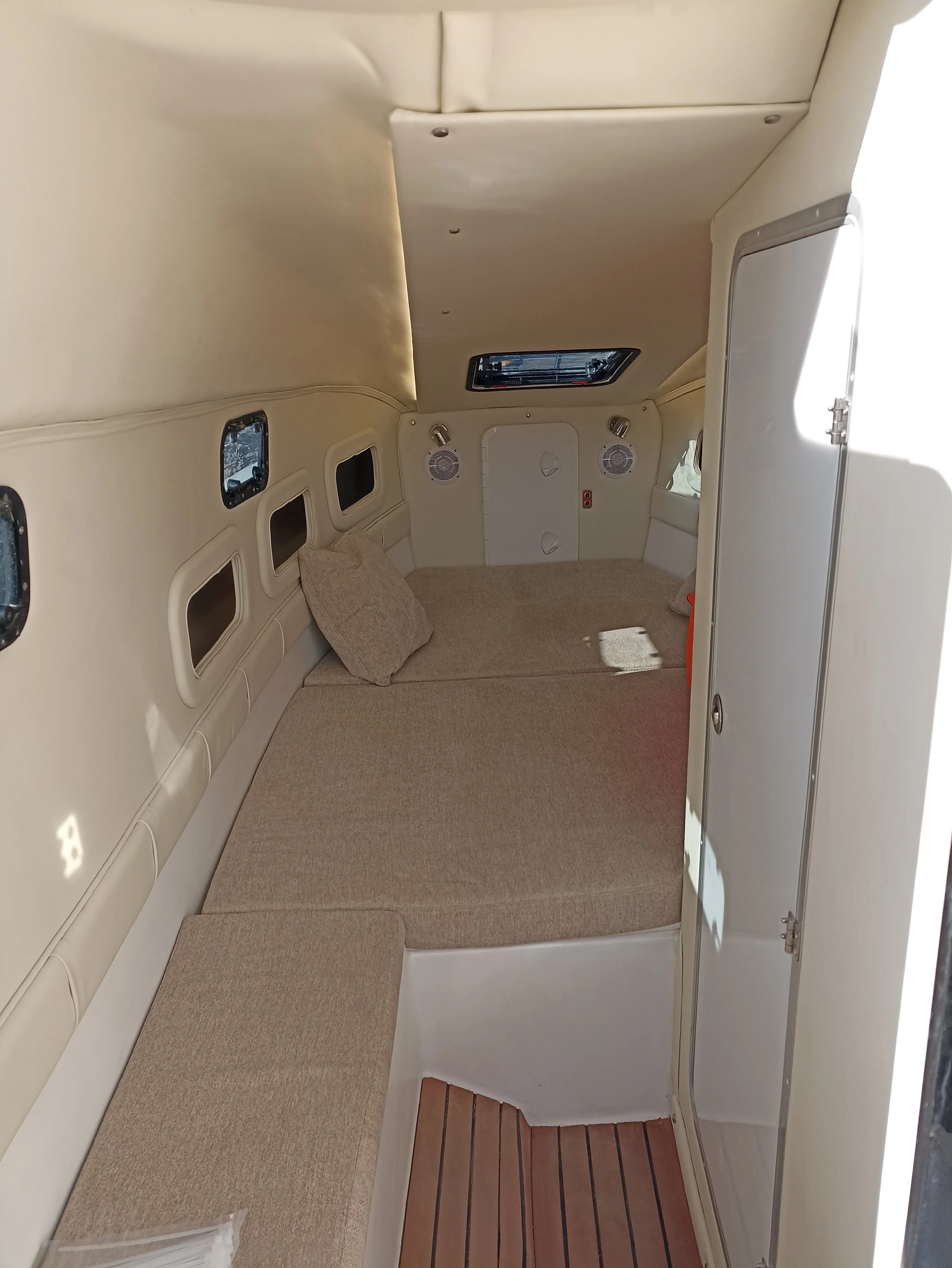 Interior of a small boat cabin with beige cushions, a pillow, and small windows on the wall.