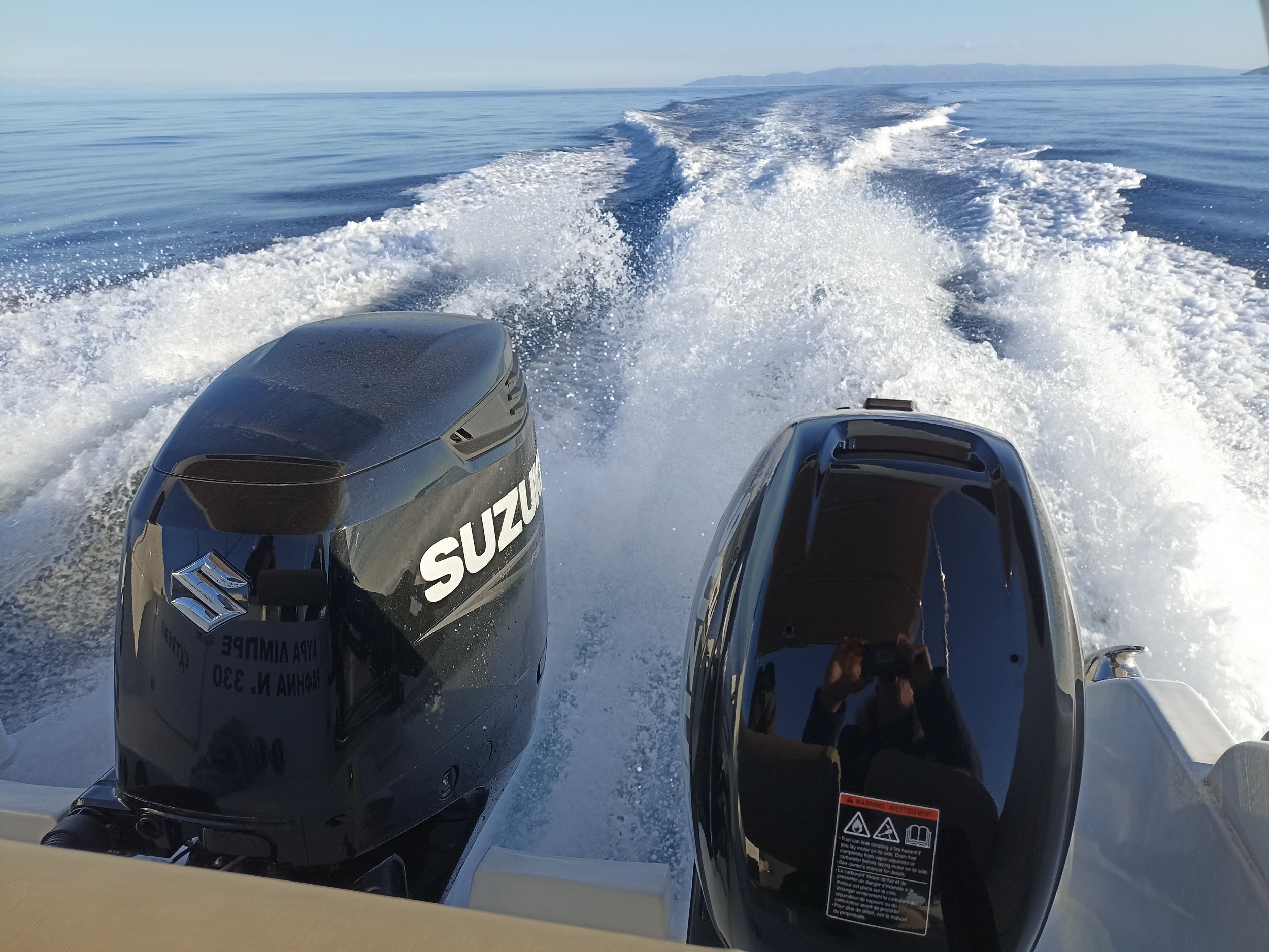 Photo taken from a boat showing two outboard motors, one labeled Suzuki, cutting through open water with a distant shoreline and mountains in the background.