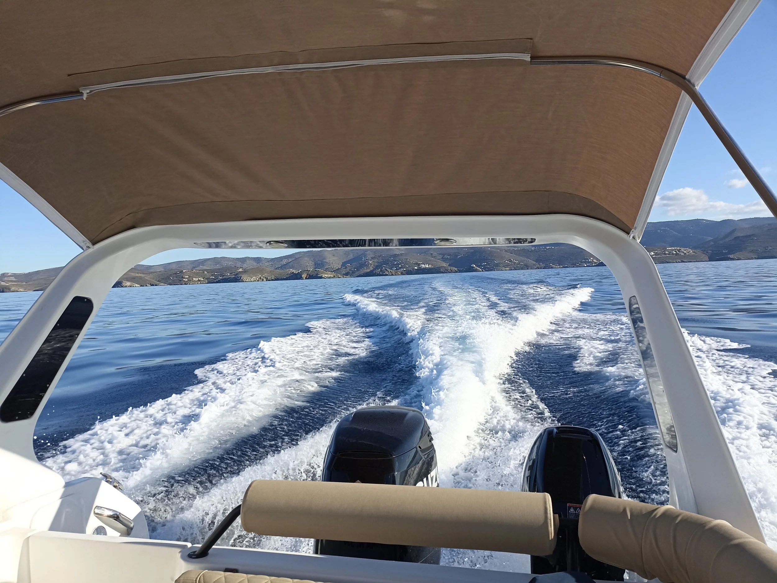 View from a boat showing two outboard motors, water trail, and distant hills on a sunny day.