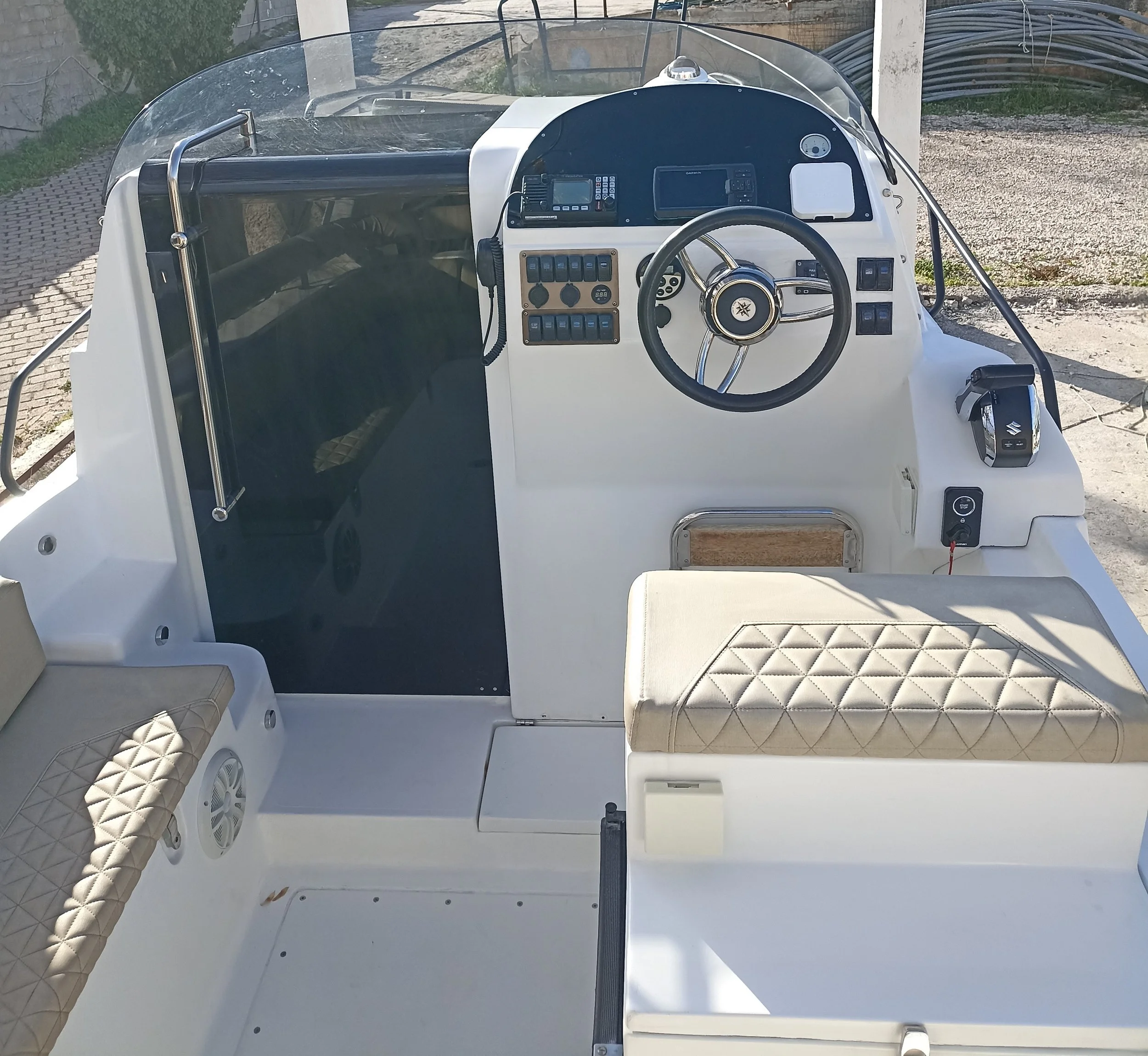 The dashboard of a small boat with navigation and communication equipment, a steering wheel, and cushioned seating areas, outdoors near a body of water.