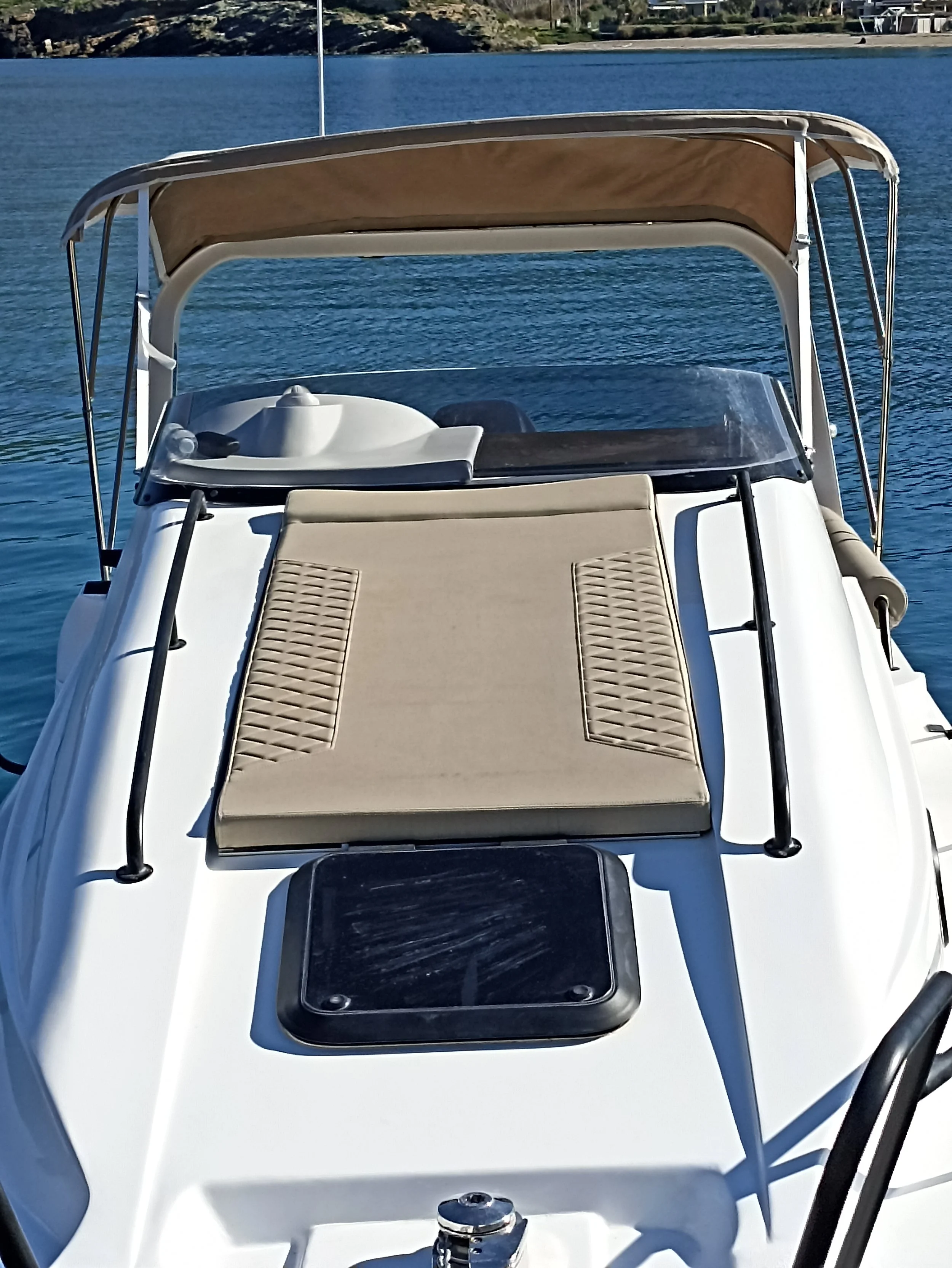 View of a white motorboat on the water with a beige sunbed and canopy, and a black hatch on the deck.