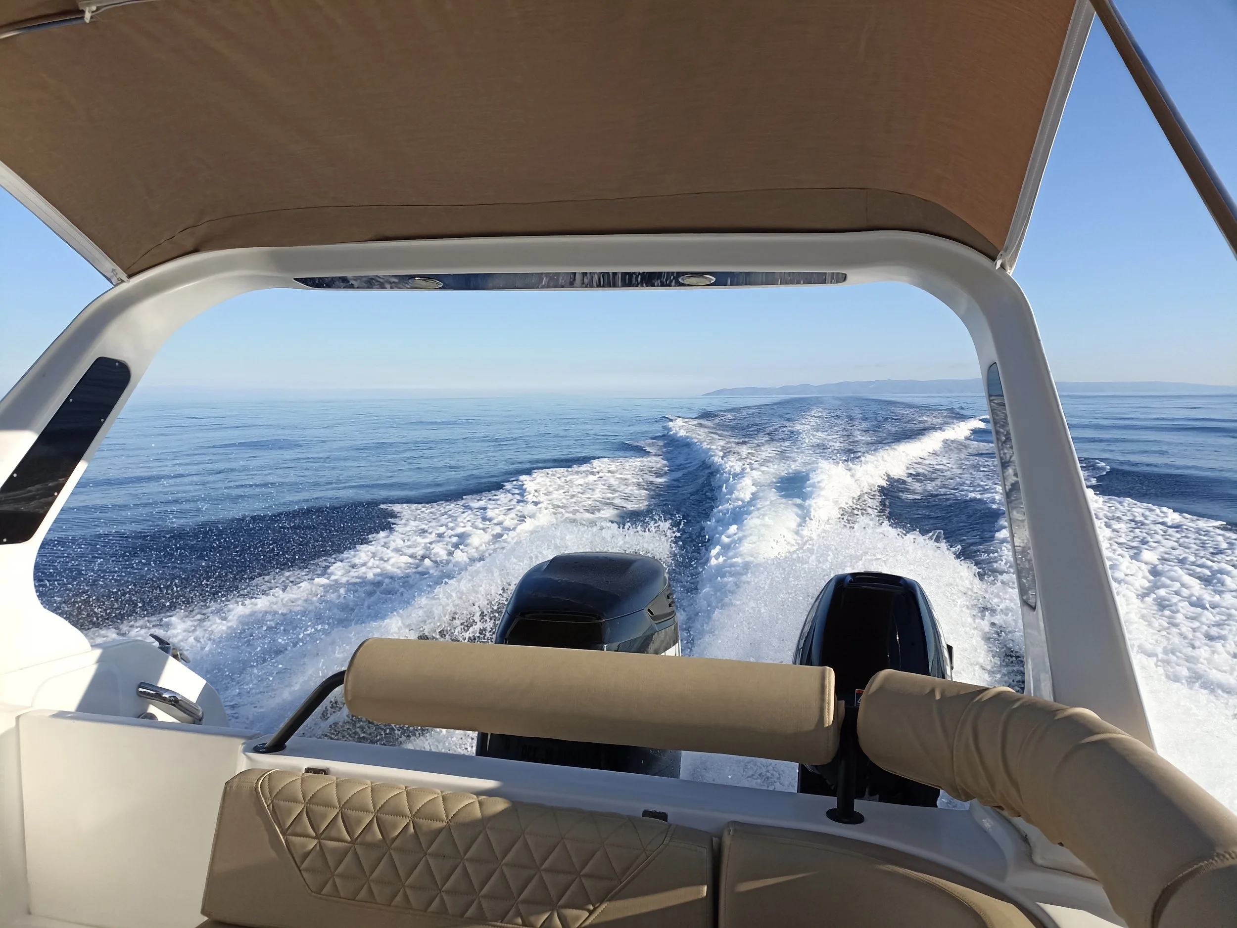 View from the back of a boat showing the open sea and wake behind the boat, with blue sky and distant land.