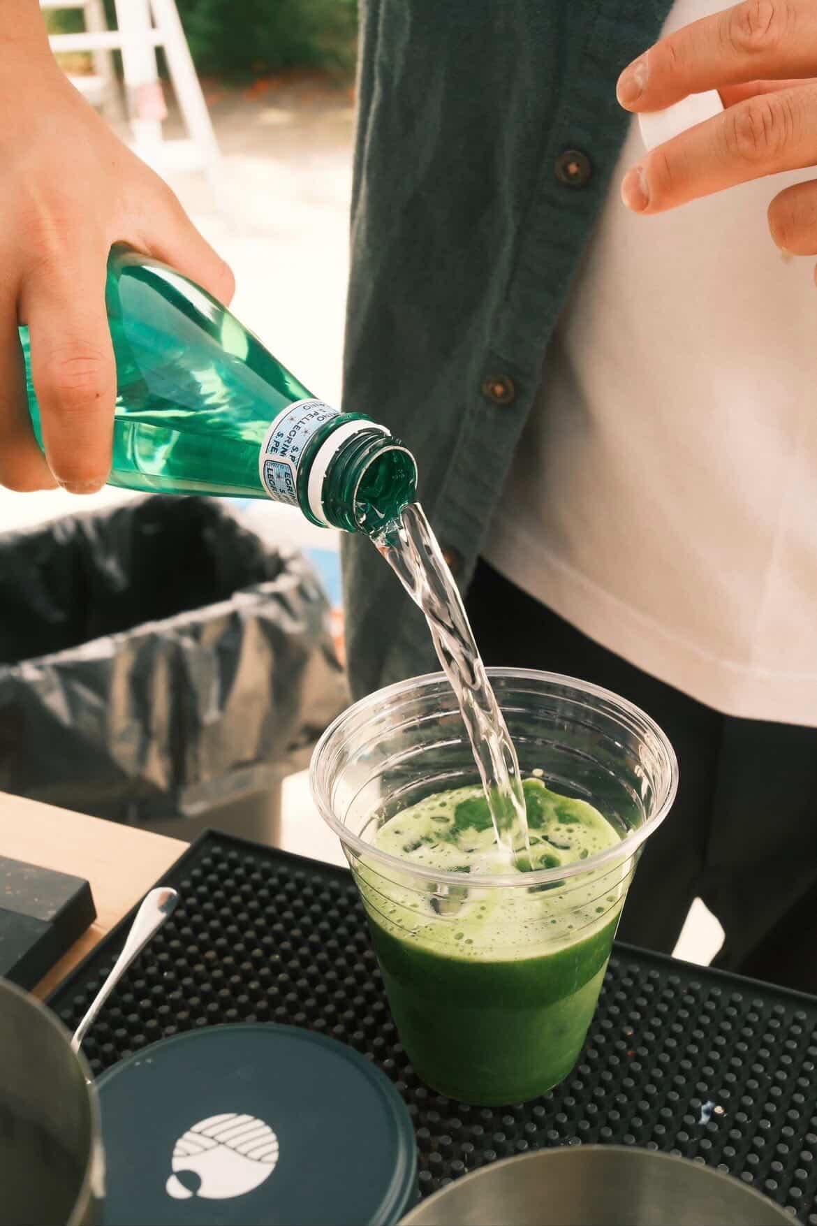 A person pouring green liquid from a green bottle into a clear plastic cup with a matcha drink inside.