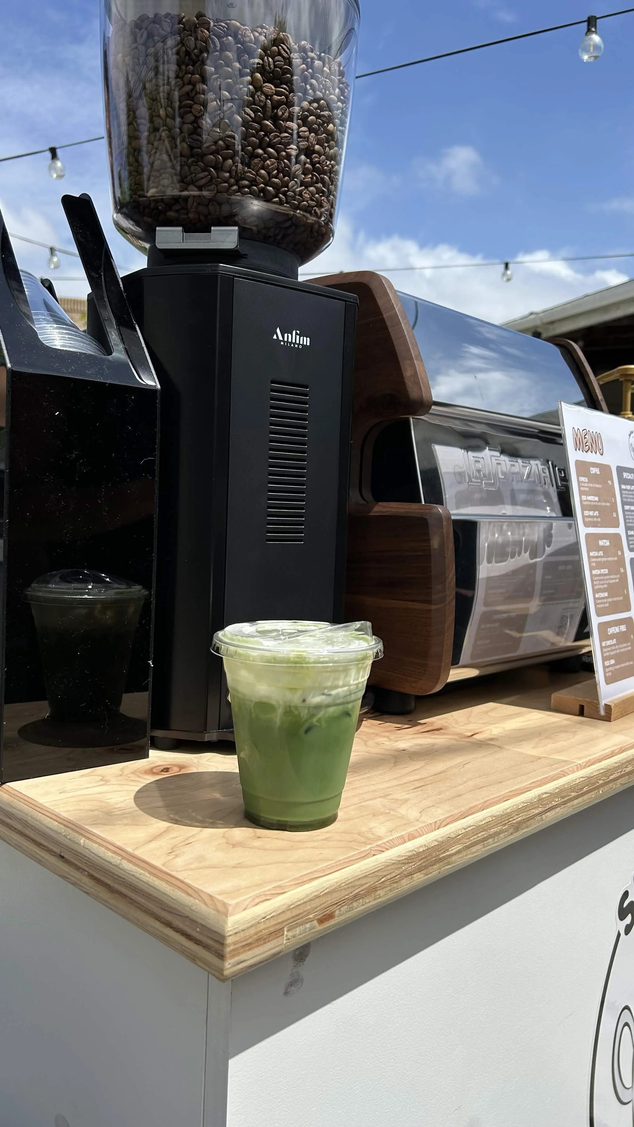 A coffee bean grinder with a large transparent container filled with coffee beans, a black coffee machine, and a menu sign on a wooden counter outdoors under string lights and a blue sky.