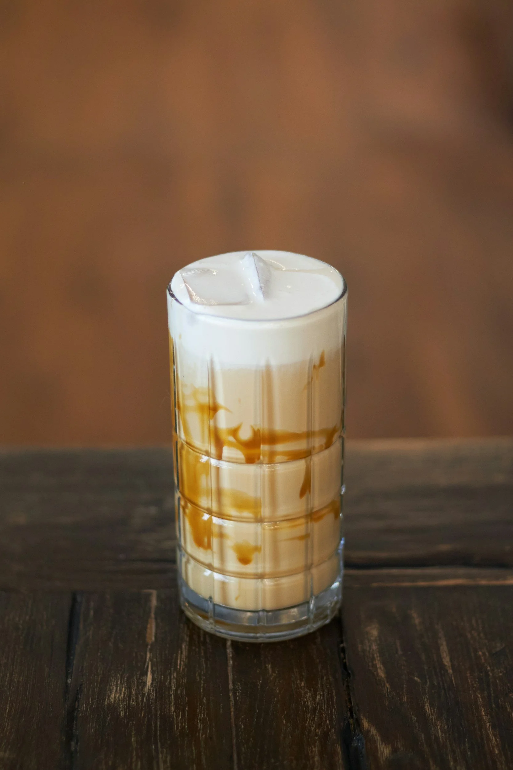 A tall glass filled with iced coffee, garnished with cream and caramel syrup, placed on a dark wooden surface.