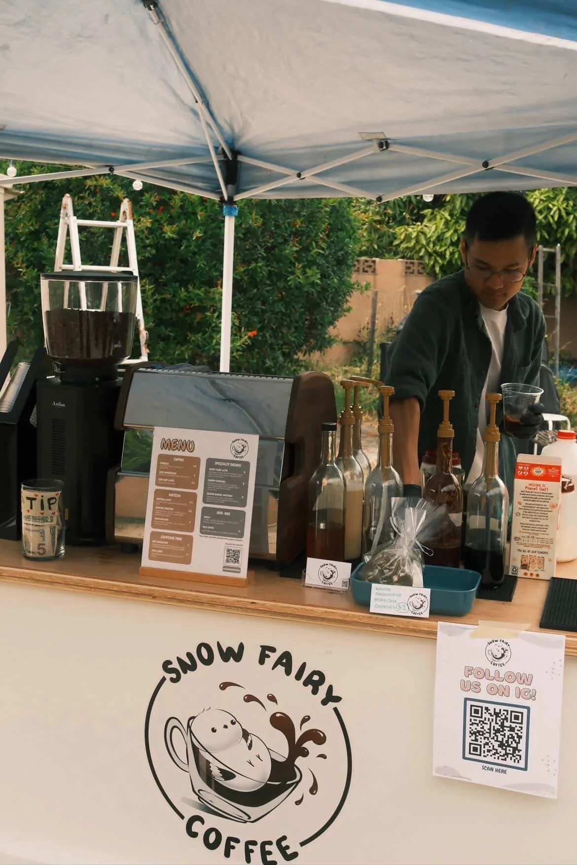 A barista behind a coffee stand at Snow Fairy Coffee, with bottled syrups, a coffee machine, and a menu. There is a sign with a QR code to follow on Instagram, and a cup with a tip jar. A tent is overhead, and greenery is in the background.