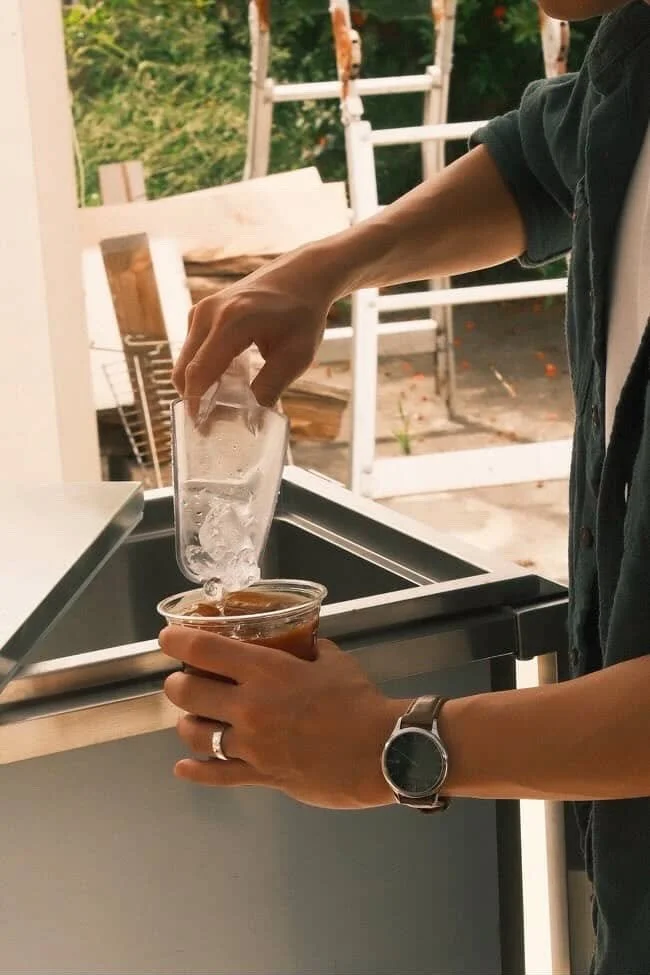 Person pouring ice water from a glass into a plastic cup with iced coffee outside next to a black countertop, with a ladder and trees in the background.