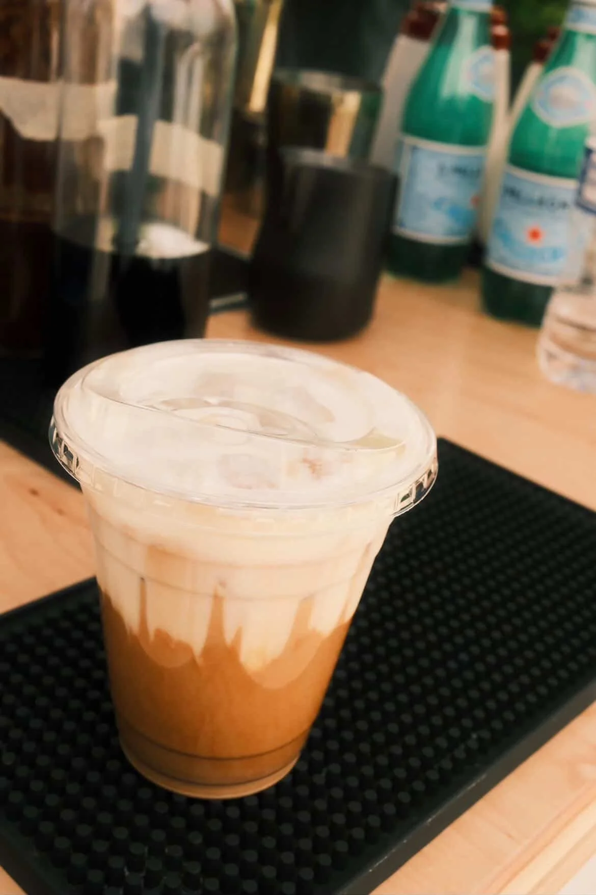 A plastic cup with a lid containing a layered iced coffee drink, placed on a black textured bar mat on a wooden counter, with bottles and containers in the background.