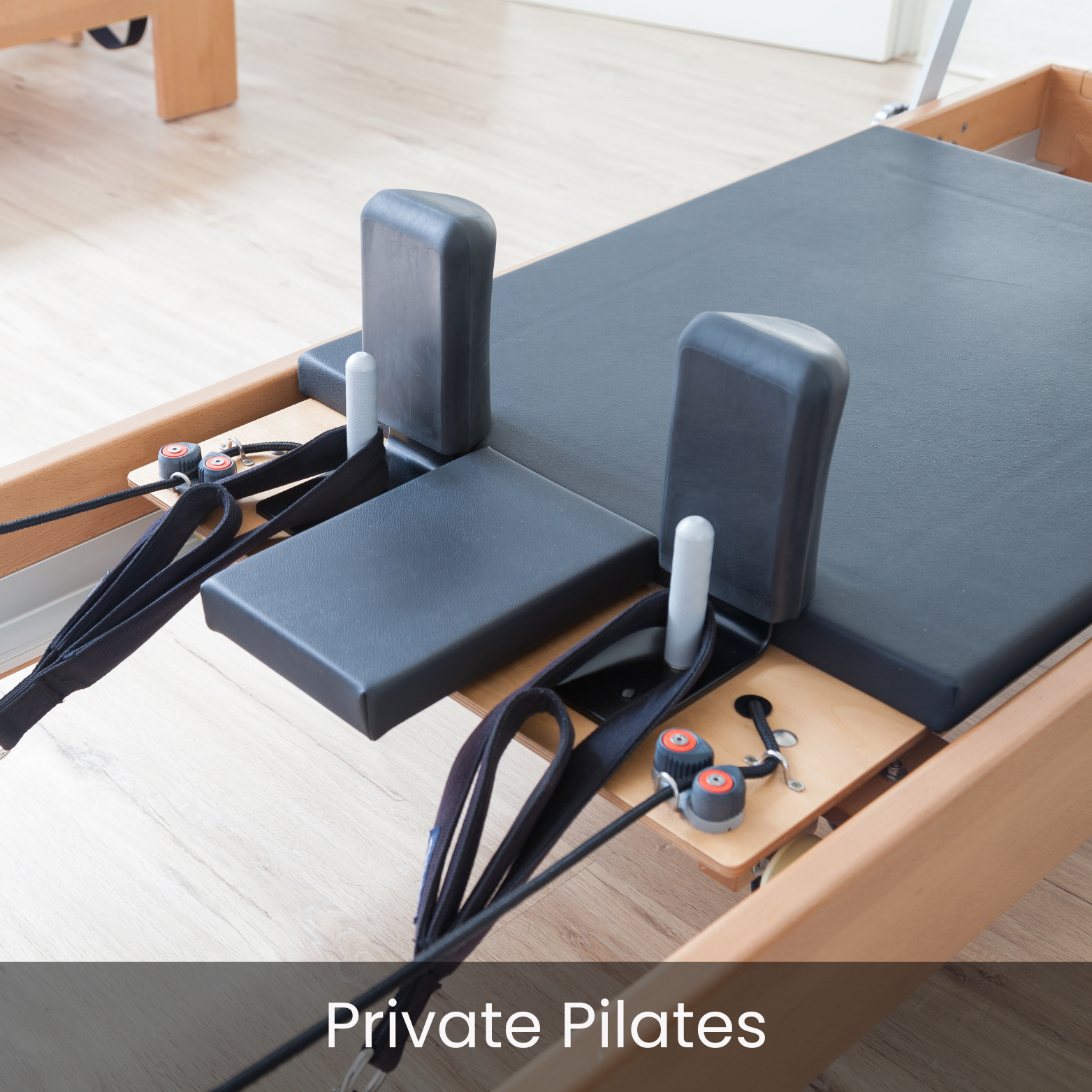 A Pilates reformer machine with black padded surfaces, white handles, and resistance cords, situated on a wooden floor in a bright exercise room.