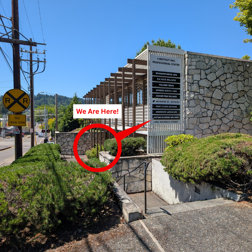 Sign for Chestnut Hill Professional Center, Autumn Blossom, with a red circle and arrow pointing to a spot with a caption 'We Are Here!' next to a stone wall and bushes, near a street with railroad crossing signals and power lines.
