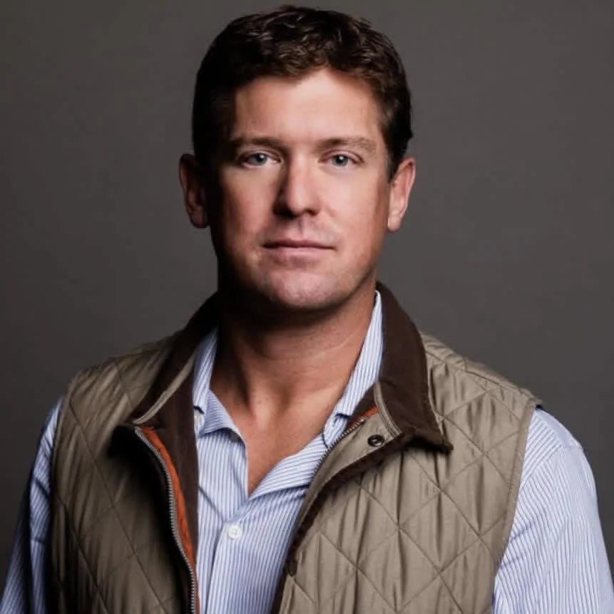 A man with short brown hair and blue eyes wearing a light blue striped shirt and a tan quilted vest against a plain gray background.