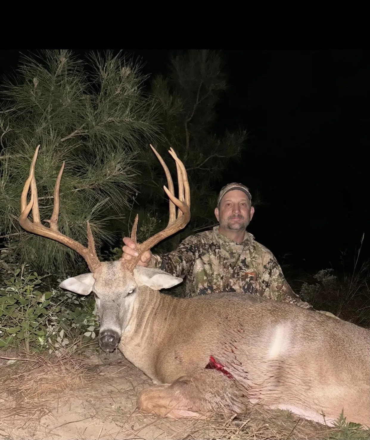 A man in camouflage clothing kneels at night beside a large, dead deer with antlers, holding one of the antlers. The deer is lying on the ground with a visible wound.