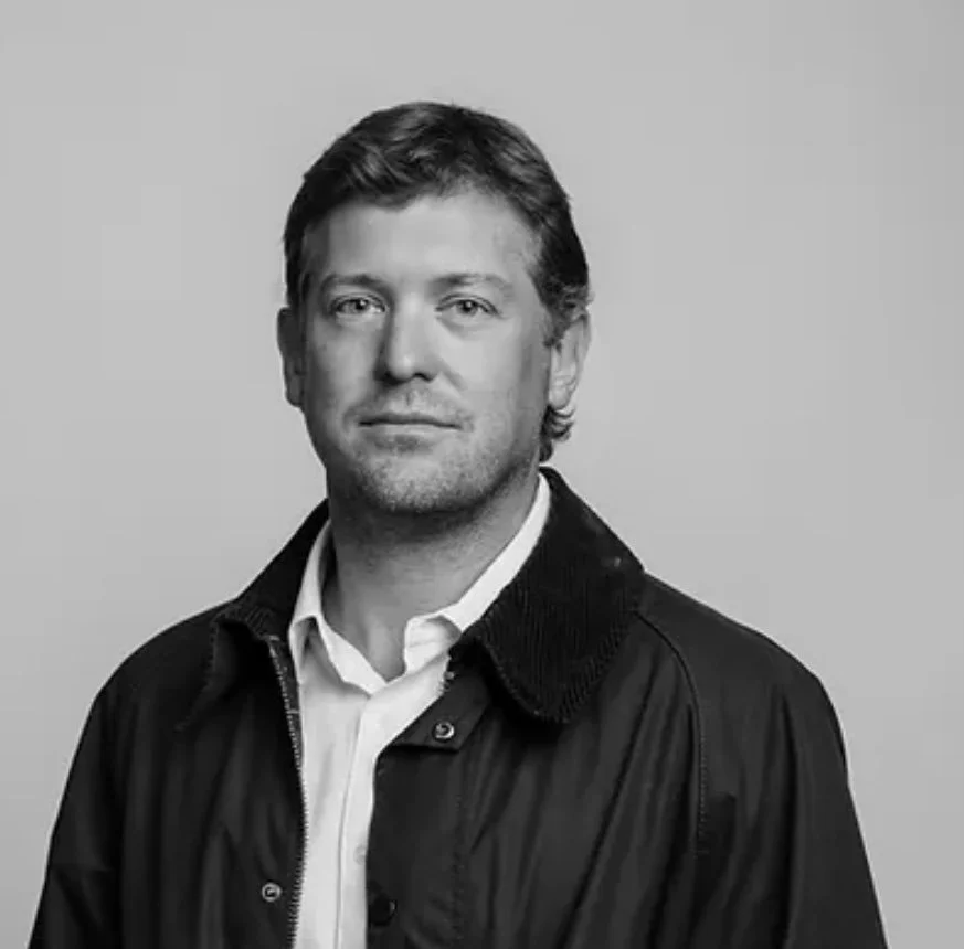 Black and white portrait of a man with neat hair, wearing a collared shirt and a dark jacket, looking at the camera with a slight smile.