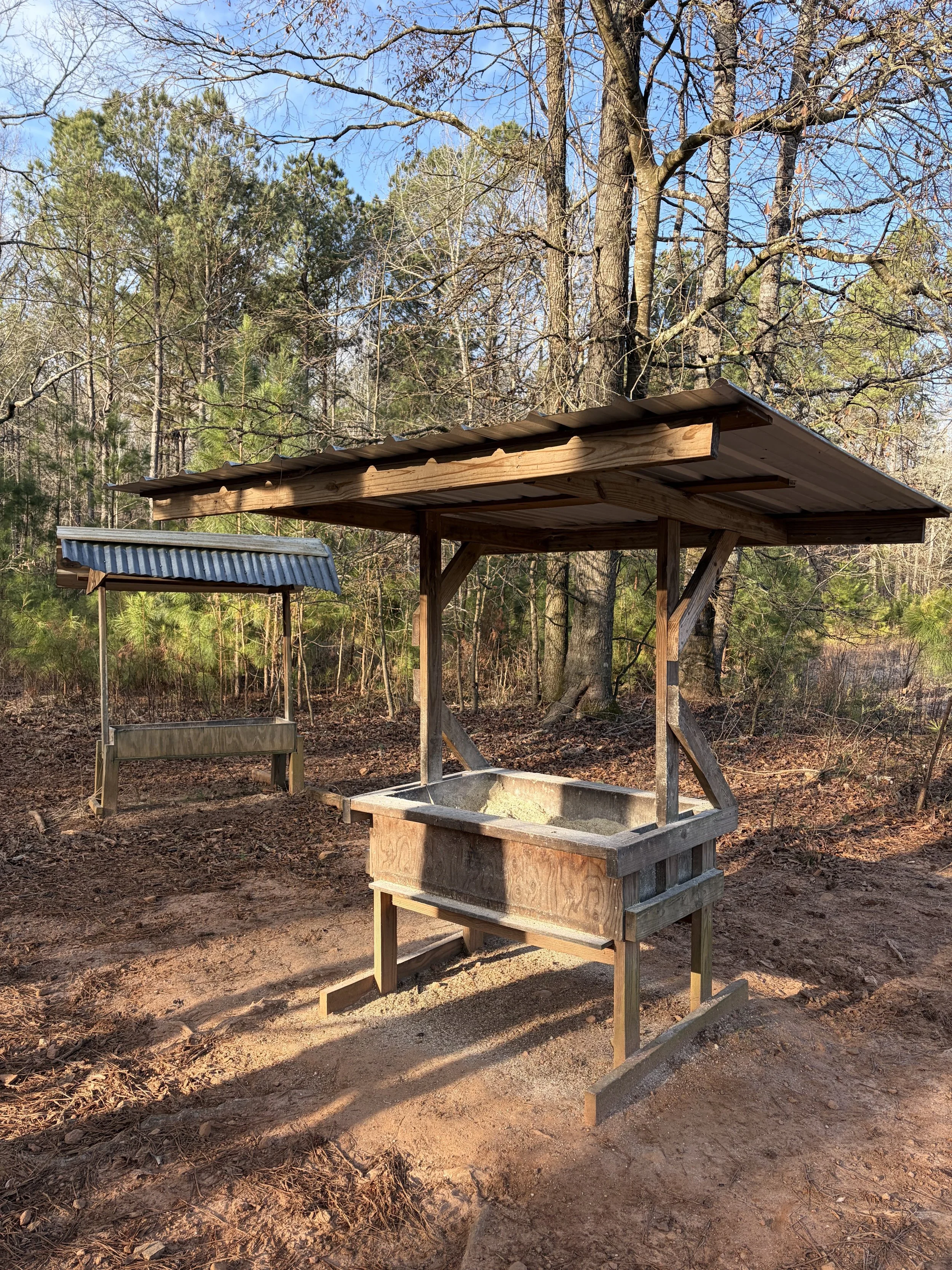 An outdoor wooden well with a corrugated metal roof and a separate shaded water trough in a wooded area with trees and a blue sky.