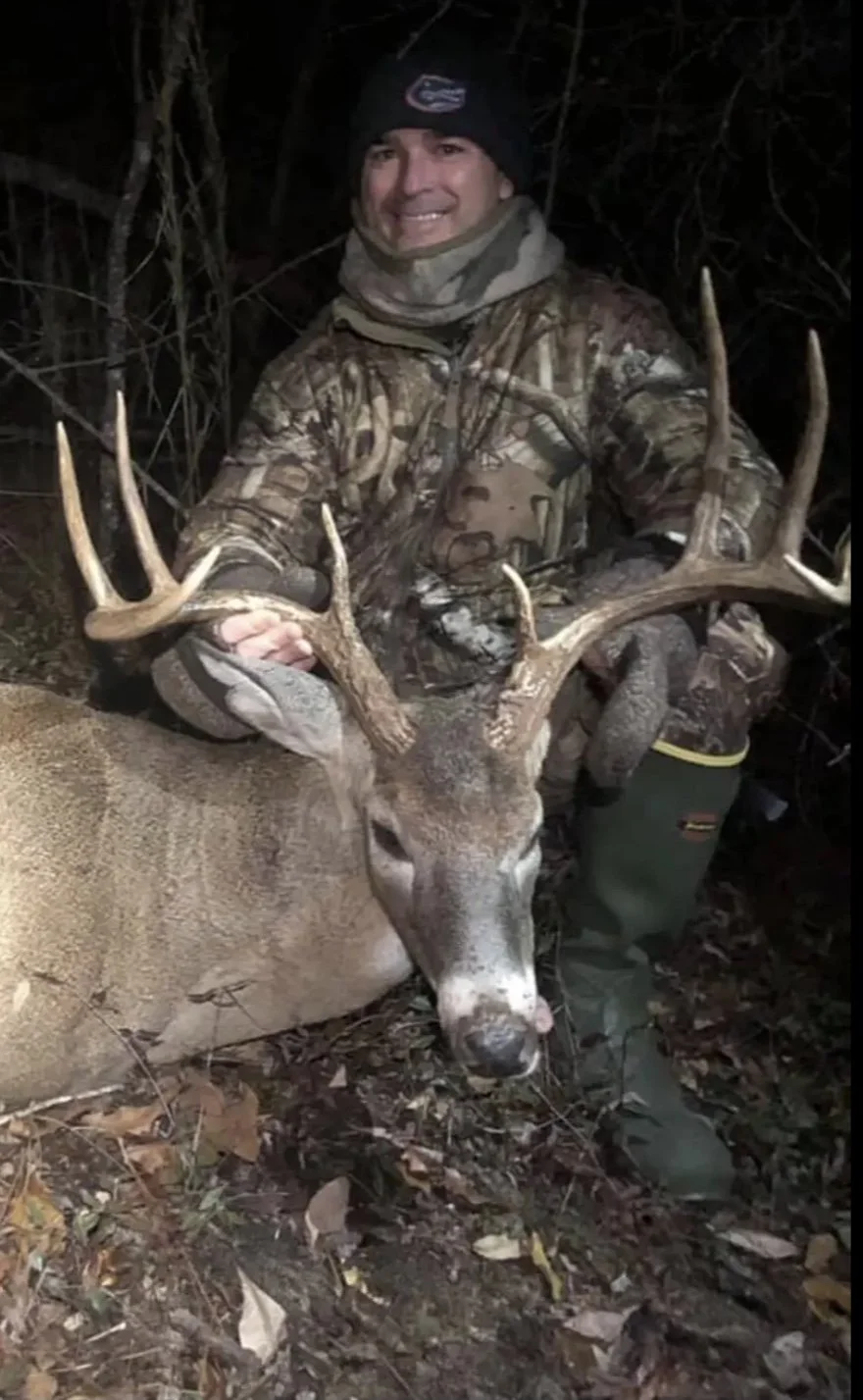 A man in camouflage clothing and a black beanie smiling and kneeling next to a large deer with antlers, holding its antlers in a wooded area at night.