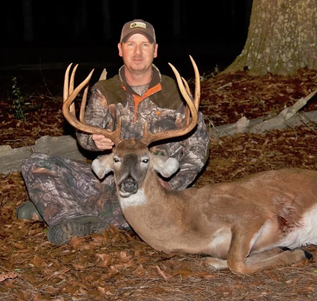A man in camouflage hunting gear kneels on the ground at night, holding the antlers of a large deer with a brown coat and prominent antlers. The scene is outdoors with trees and fallen leaves, illuminated by a flash.