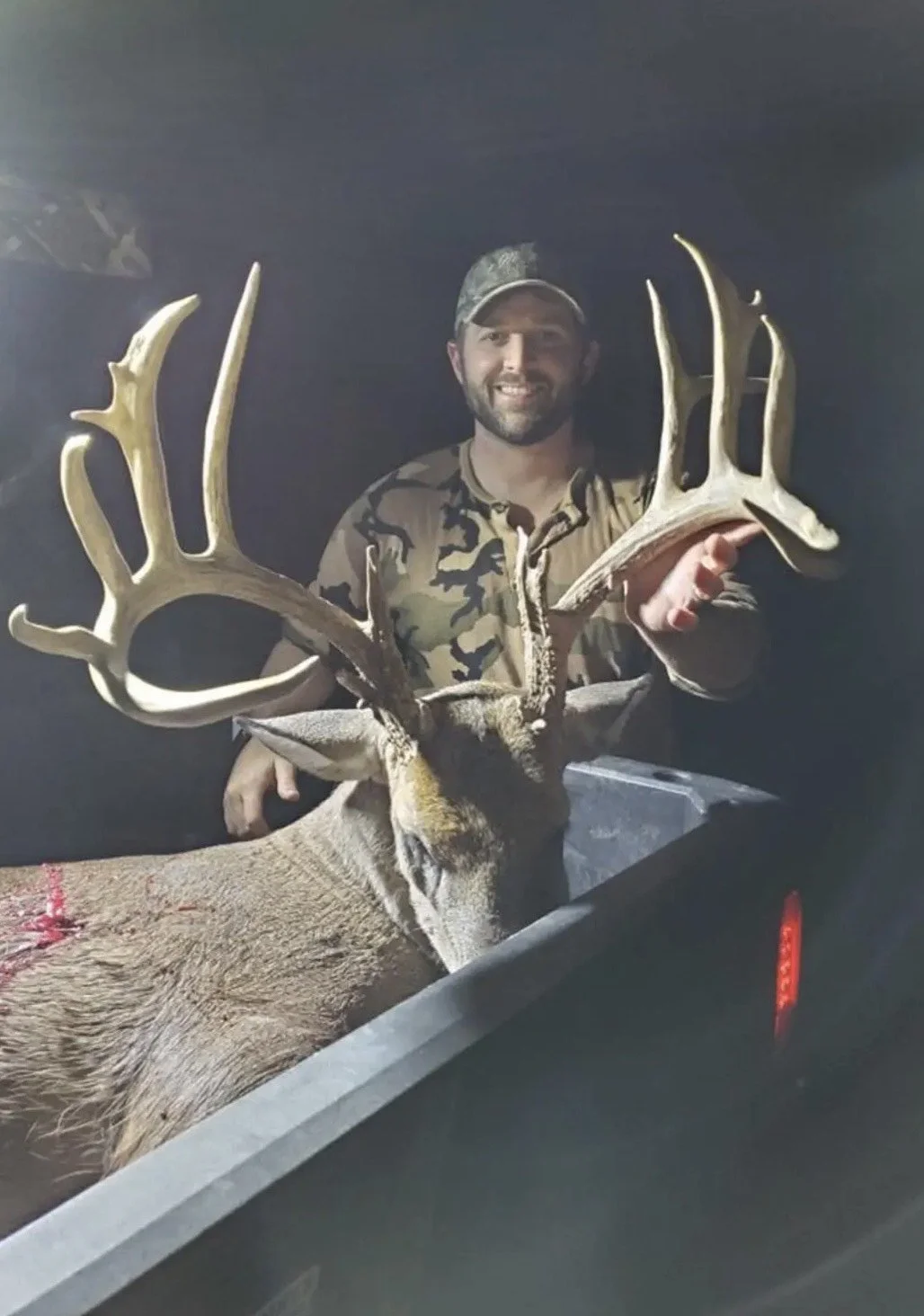 A smiling man in camouflage clothing and a cap holding a large deer with impressive antlers in a truck bed at night.