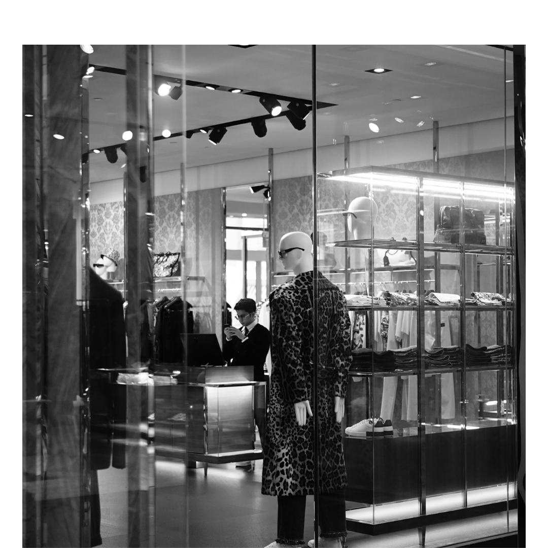 Mannequin dressed in leopard print coat and trousers displayed in a store window with clothing and accessories behind glass shelves, and a person shopping inside the store reflected in the window.