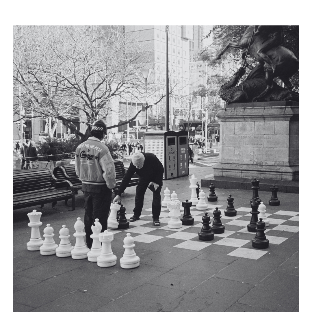 Two people playing chess outdoors on a large chessboard in a city park. One person is moving a chess piece while the other observes. There are benches nearby, and a statue of a man on a horse in the background, along with leafless trees and tall buildings.
