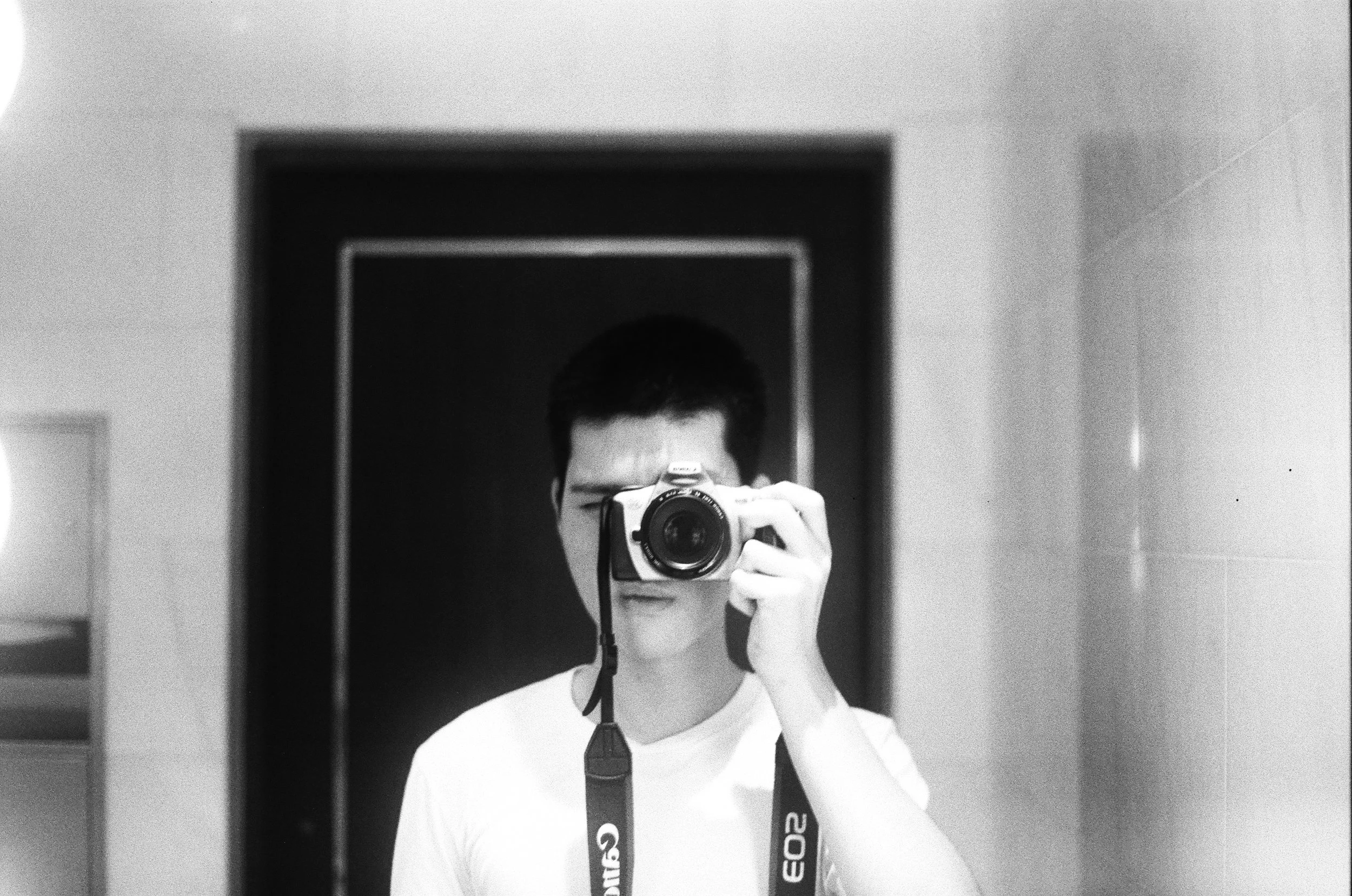 A young man taking a selfie in a mirror with a camera, with a dark background behind him and white walls to the sides.