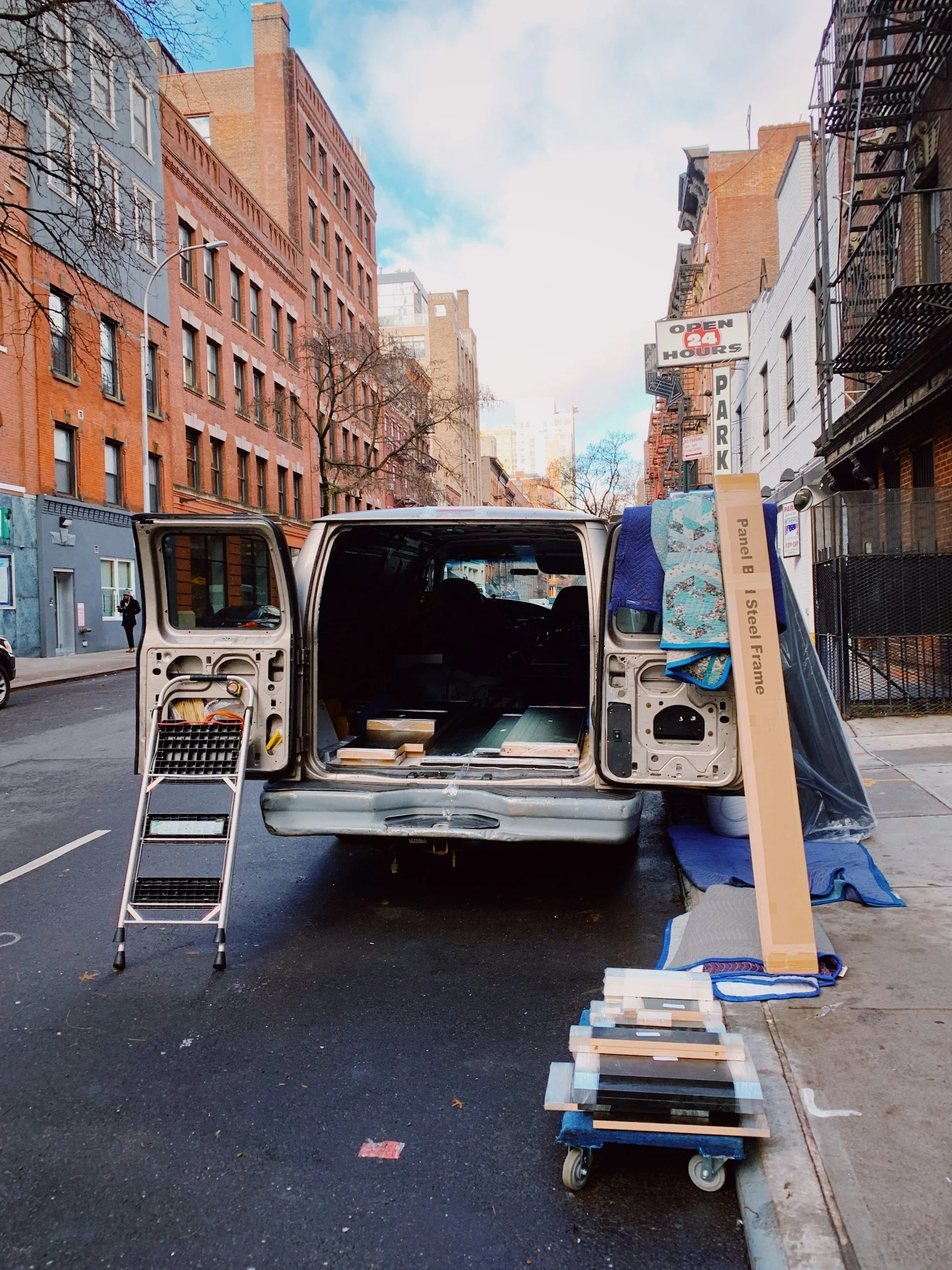 A street scene with an open van parked on the side, containing building materials, a ladder, and a shelf. There are brick buildings, a parking sign, and a blue sky with clouds in the background.