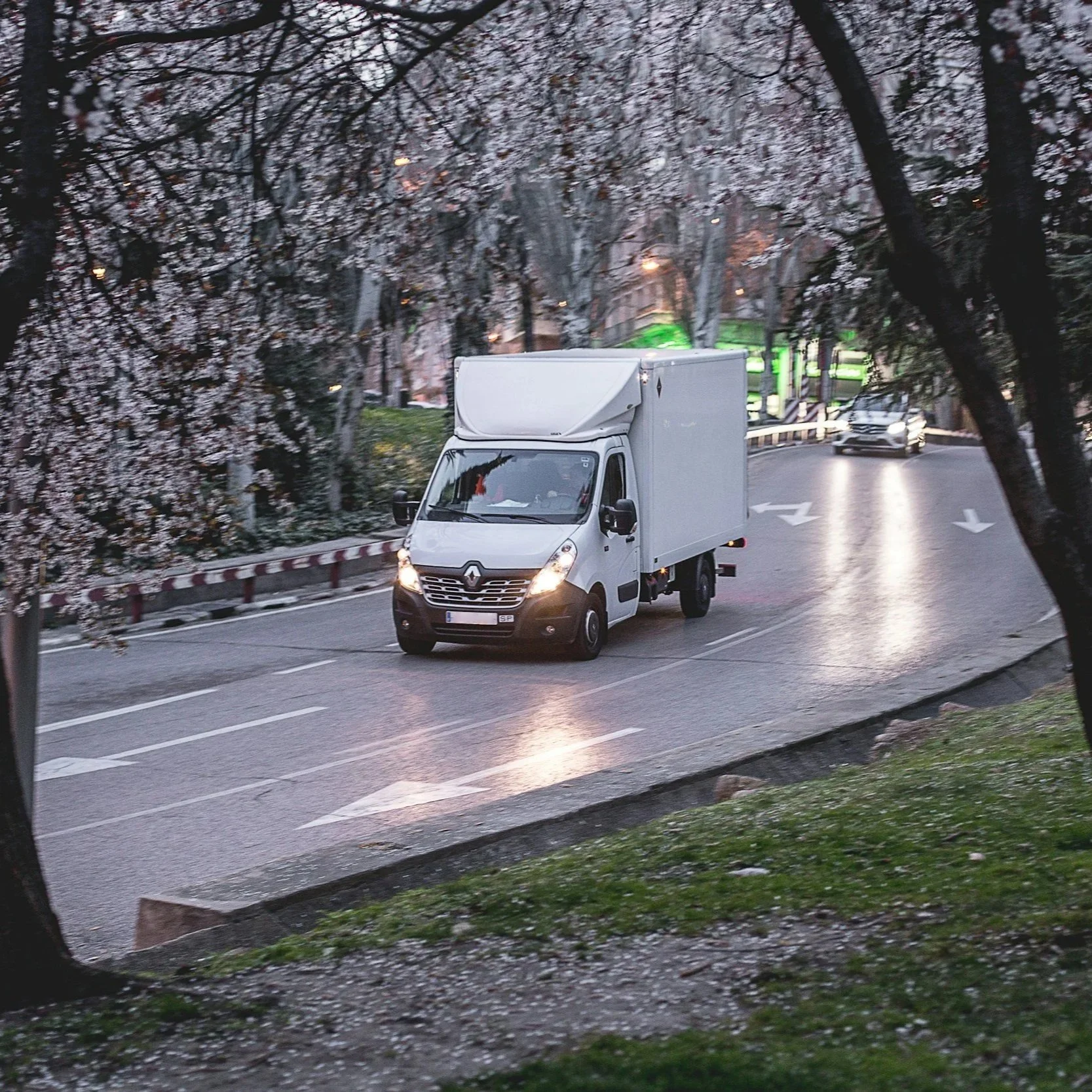 A white delivery truck driving on a wet road lined with trees in what appears to be early evening or dusk. The trees have flowers, possibly cherry blossoms, and there are cars with headlights on in the background.
