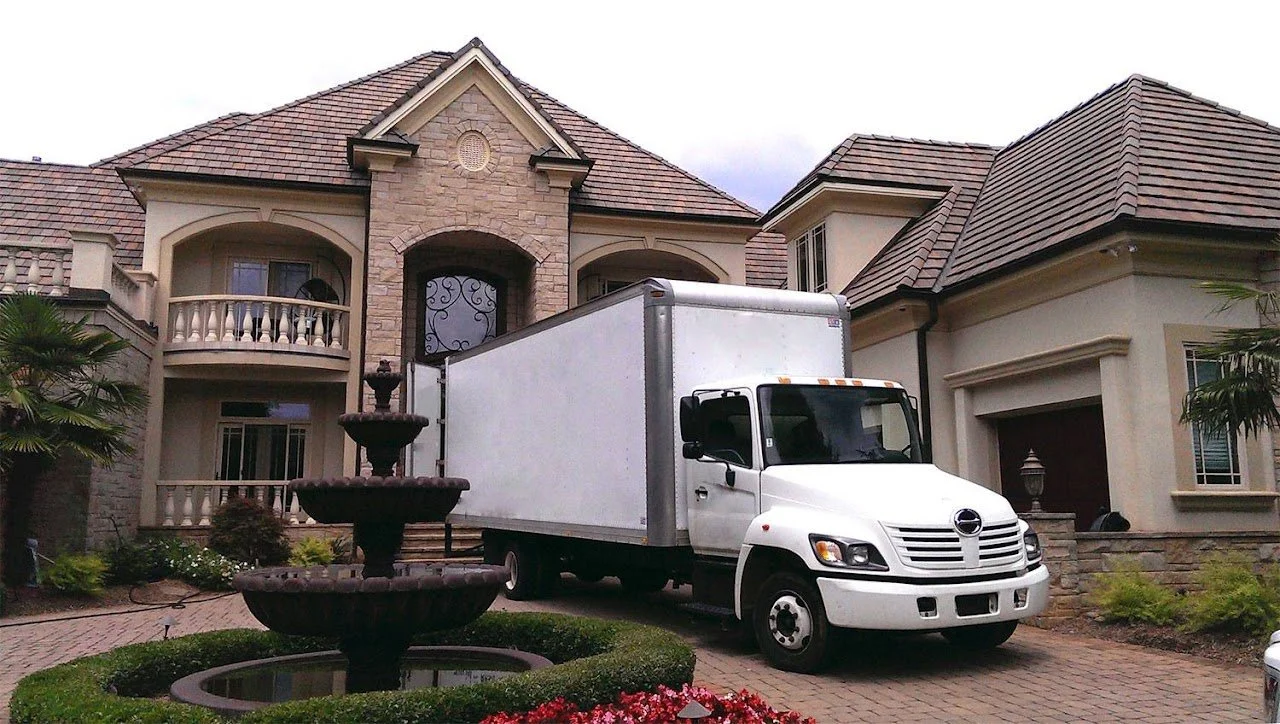 A white delivery truck parked in front of a large, luxurious house with a fountain and landscaped yard.