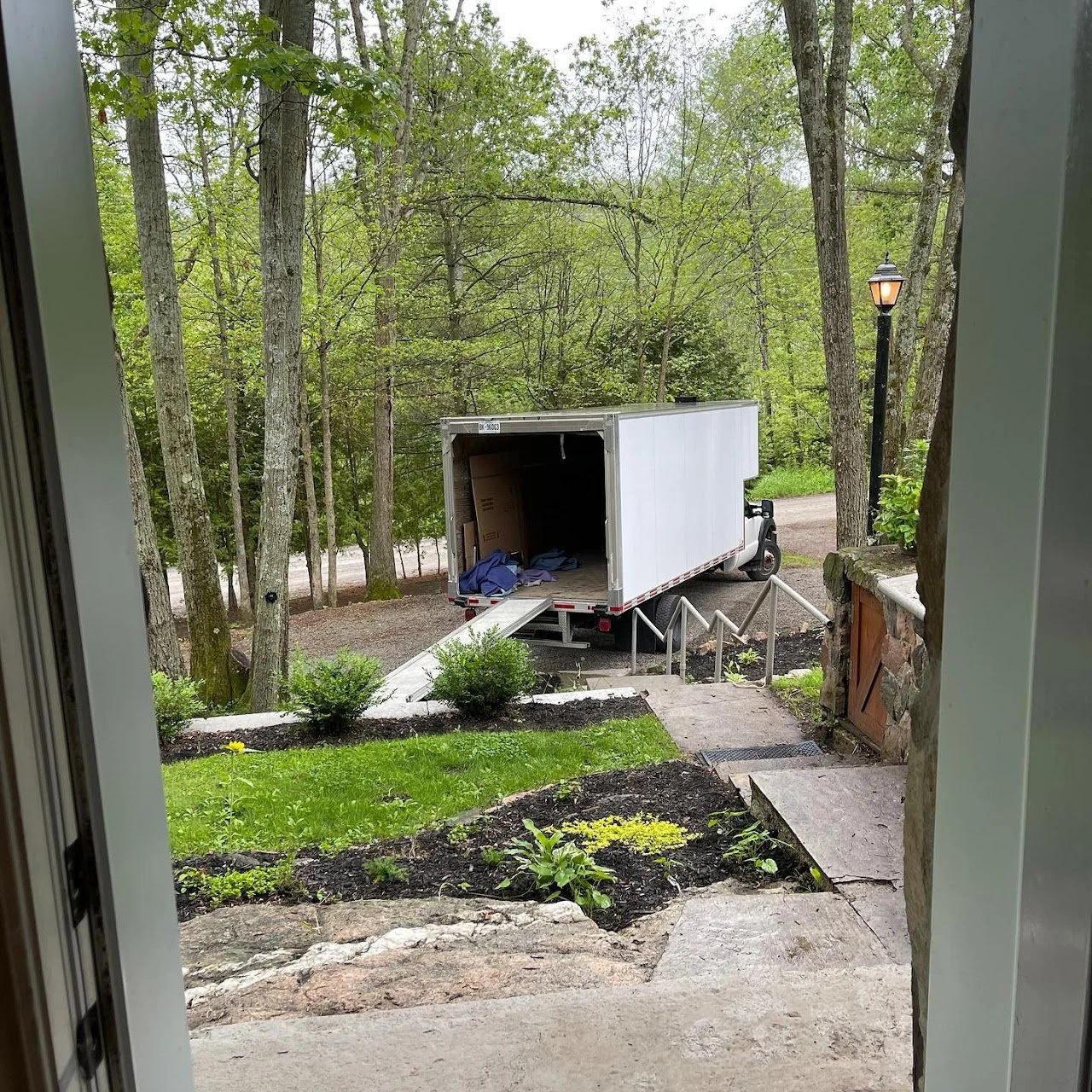View from a doorway showing an open moving truck parked on a gravel driveway, with a ramp leading into the truck. There is a set of stairs with a handrail, some green bushes, and trees with lush green leaves in the background. A lamp post is also visible.