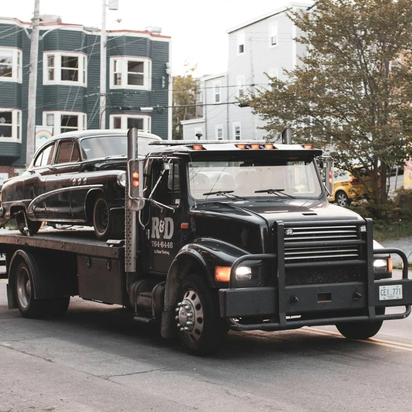 A black tow truck with the logo 'R&D' is transporting a vintage black car on its flatbed.