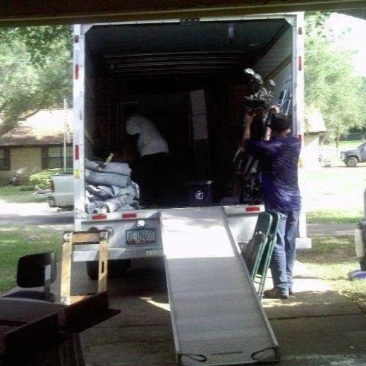People loading items into a moving truck with a camera operator filming the scene.