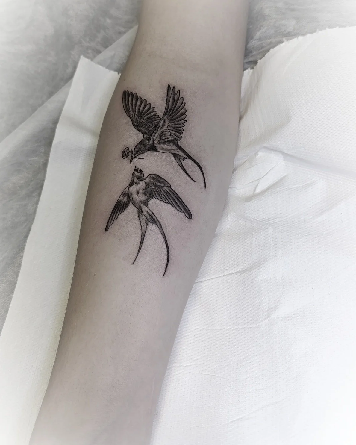Juodai pilka dviejų skrendančių paukščių tatuiruotė ant dilbio, vienas snape neša mažą gėlę.