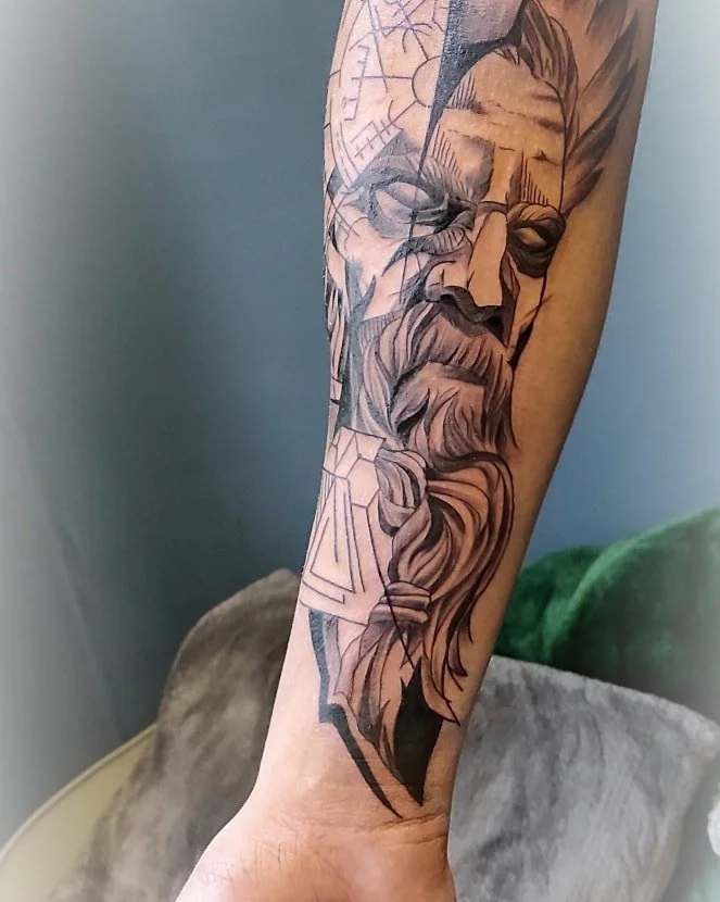 Linijinio stiliaus (line art) barzdoto vyro ilgais plaukais ir su akiniais veido tatuiruotė ant dilbio.