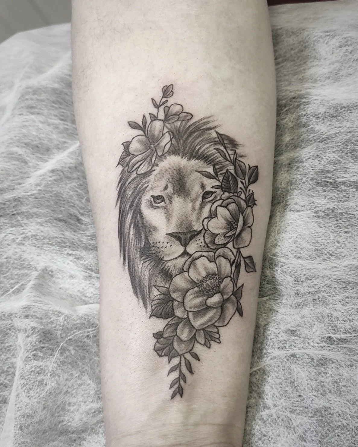 Liūto snukio tatuiruotė ant rankos, apsupta detalių gėlių ir lapų.