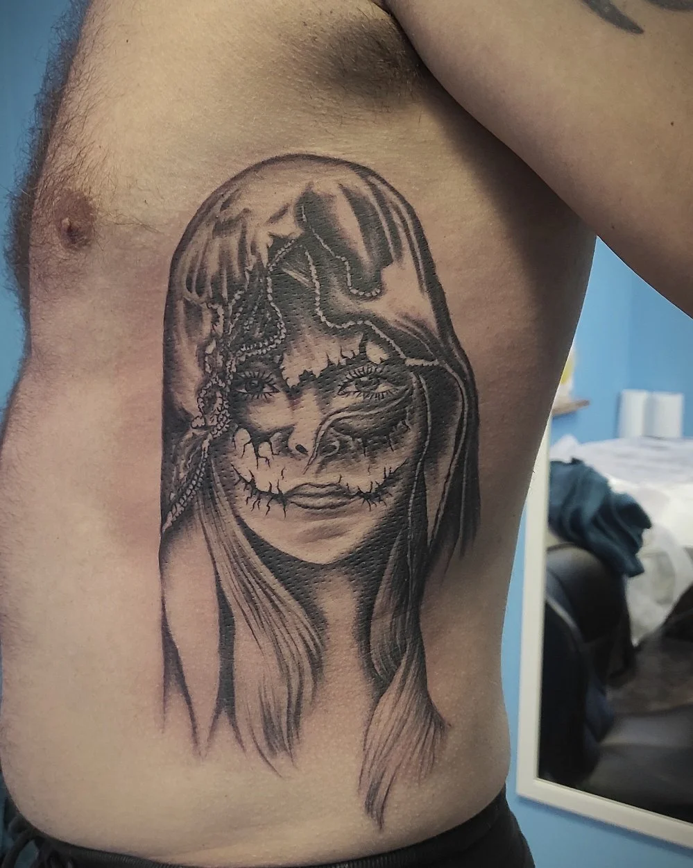 Moters veido tatuiruotė ant šonkaulių, kurios pusė pavaizduota kaip kaukolė su veido dažais.