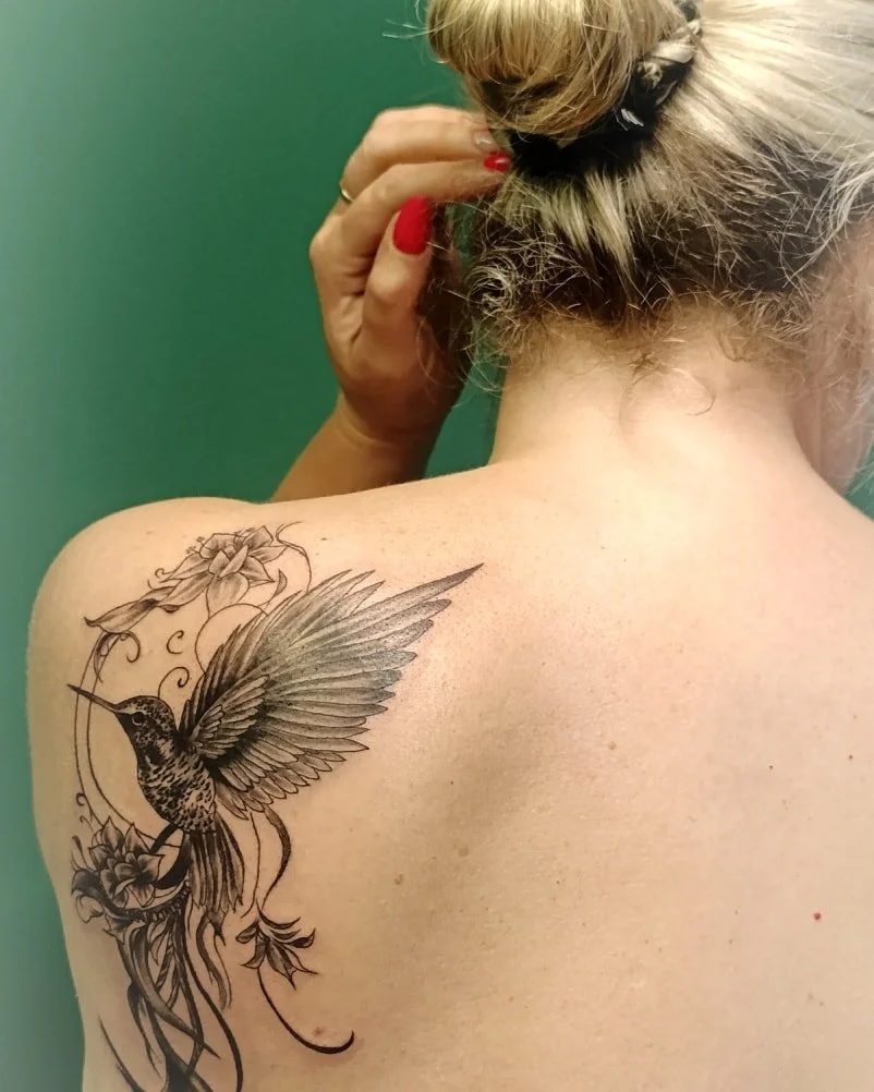 Gėlių apsupta kolibrio tatuiruotė ant moters mentės.