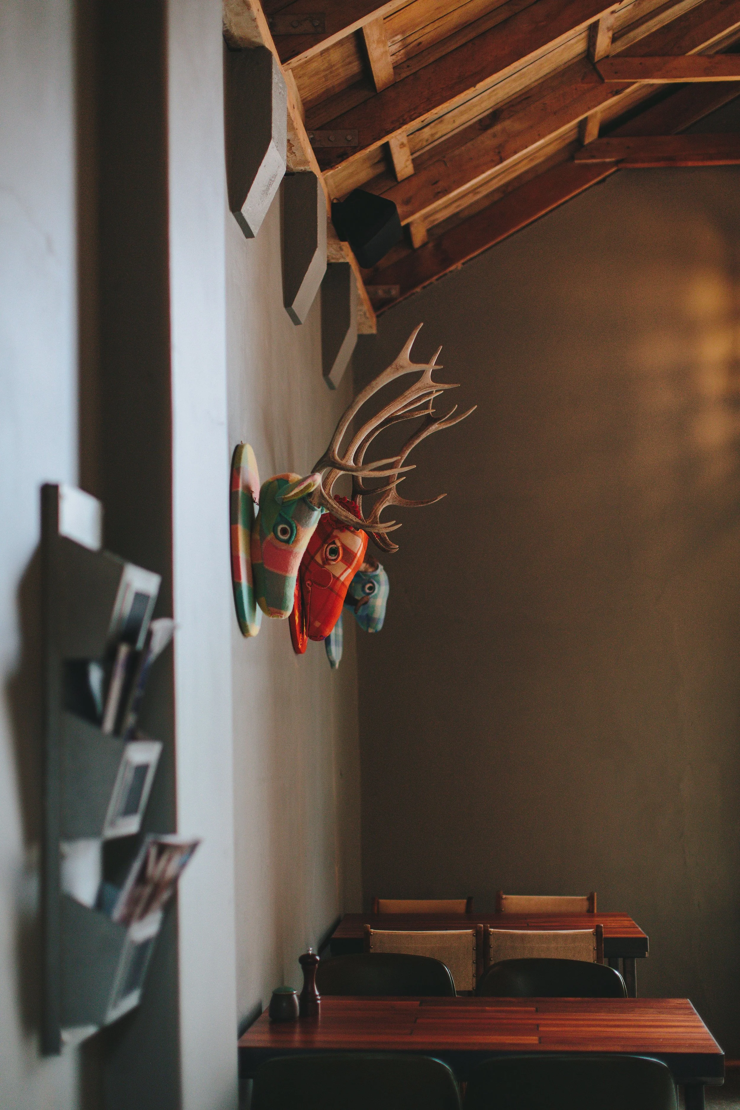 Interior of a room with wood-paneled ceiling, a gray wall with colorful deer head sculptures, and dark wood tables with chairs.