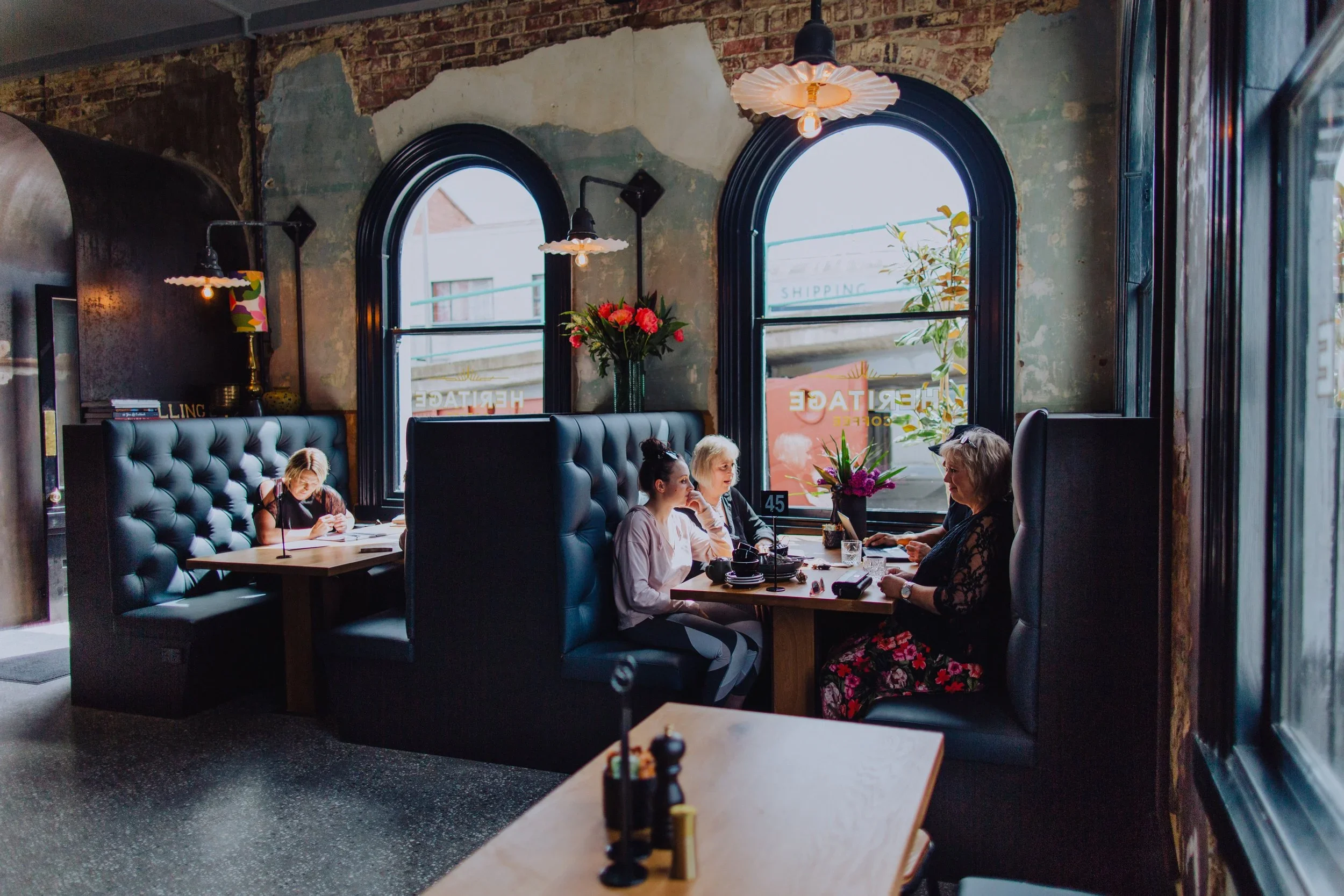 Interior of a cozy restaurant with black tufted booth seating, large arched windows with black frames, and exposed brick walls. Four women are seated at a table near the window, enjoying a meal and conversation. Decor includes colorful flowers and modern lighting.