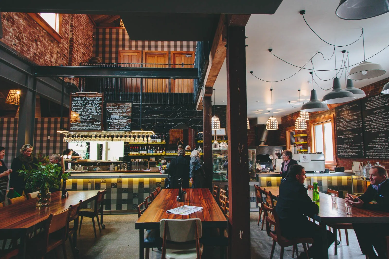 Inside a cozy cafe with wooden tables and chairs, a bar counter with black and yellow checkered design, hanging lamps, and a chalkboard menu on the wall. Several customers are seated and interacting, with some standing in line at the counter.