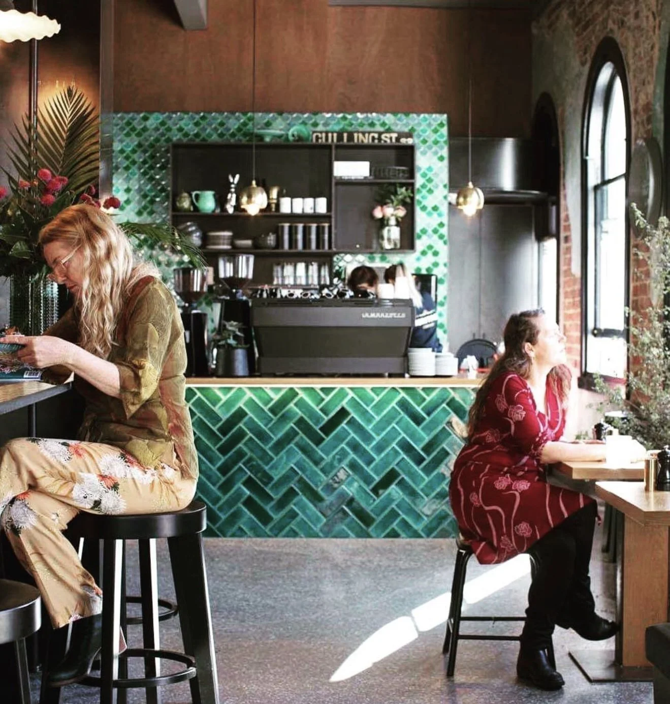 Inside a modern coffee shop with two women, one sitting at the counter looking at her phone and the other sitting at a table near a large window looking outside. The shop features green tile decor, wooden accents, and large arched windows.