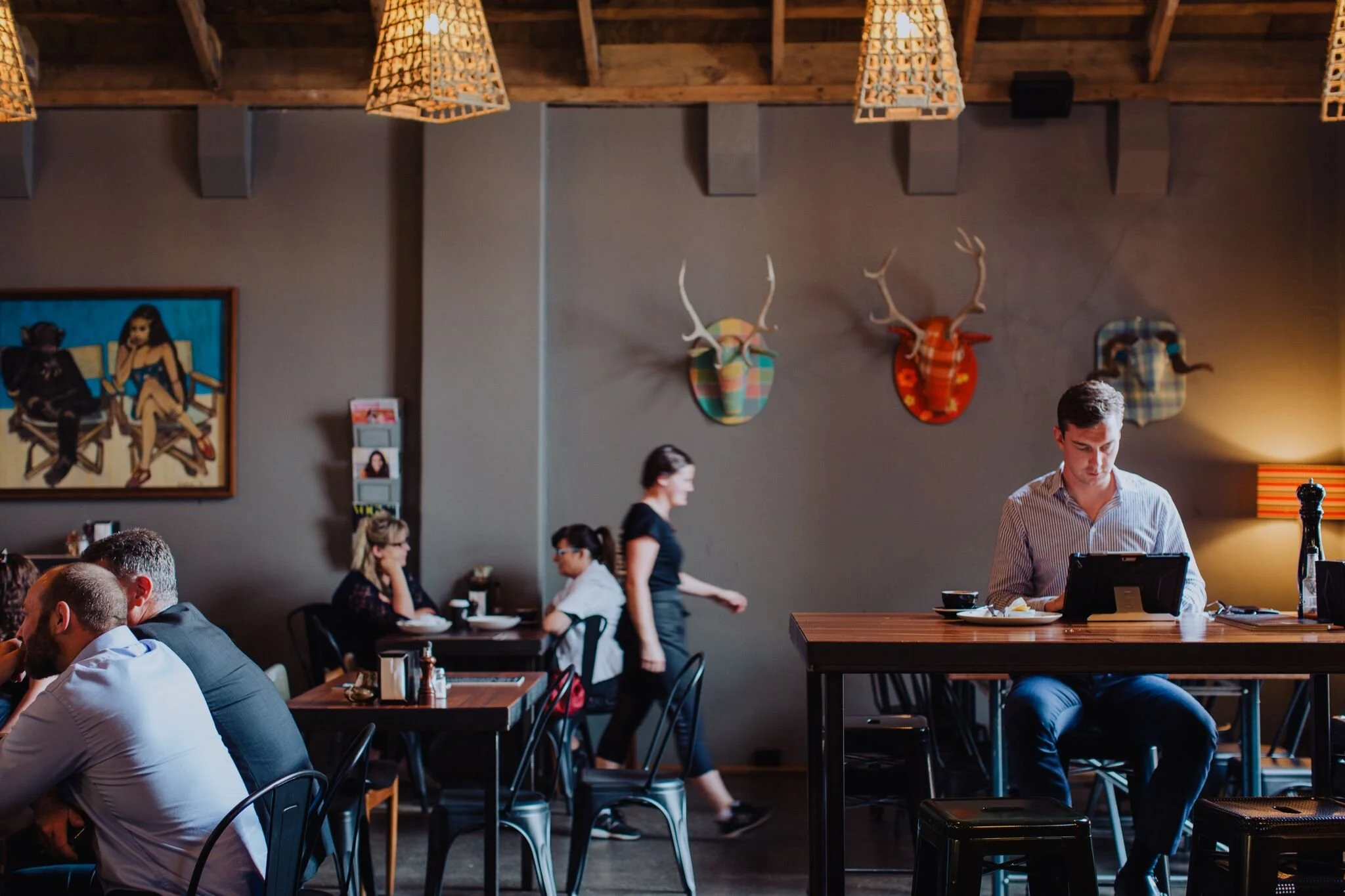People dining in a restaurant with a man working on a tablet at a high table, and a waitress walking past. Decor includes paintings and mounted antlered animal heads on the wall.