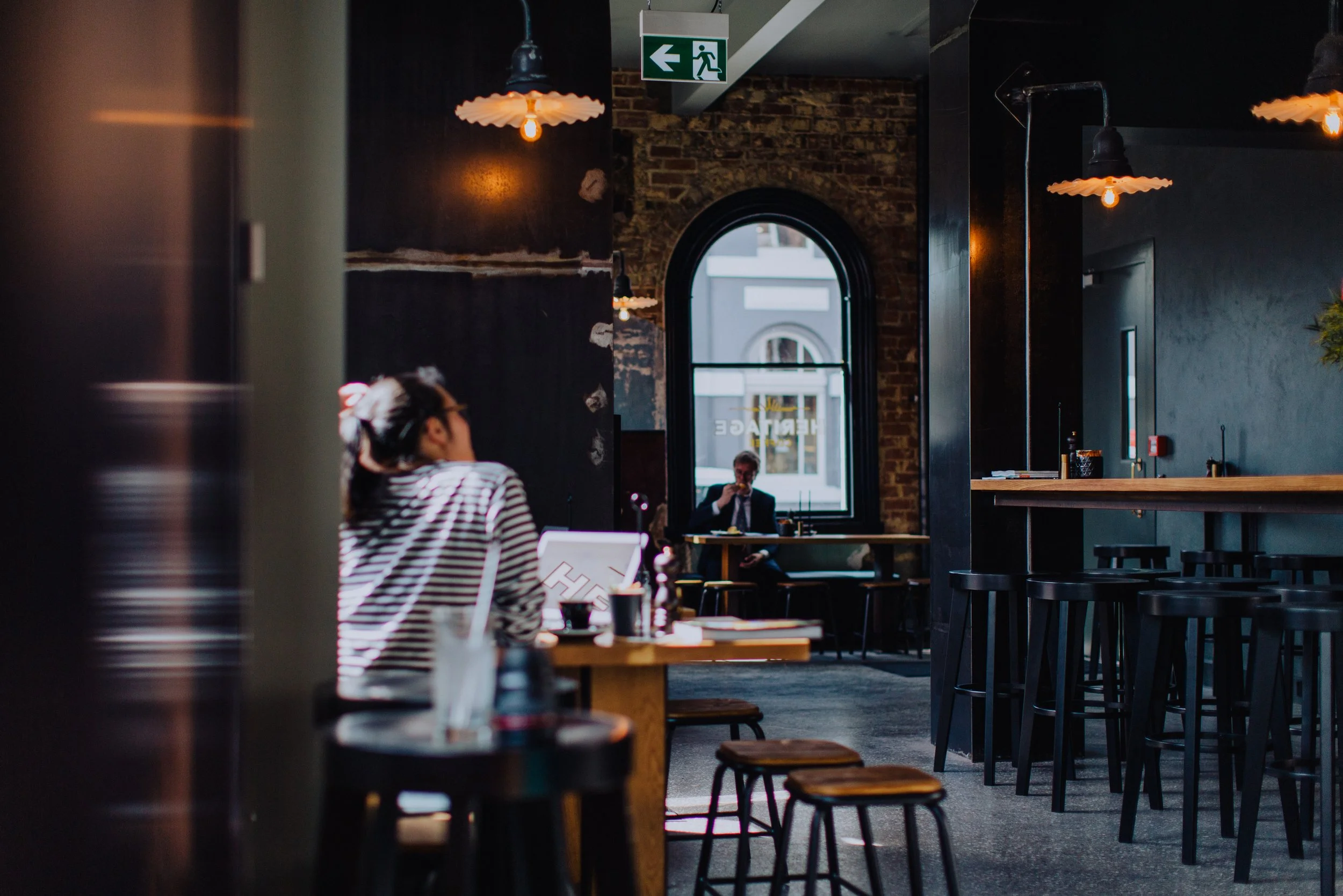 Interior of a modern coffee shop with black walls, wooden tables, and stools. A woman with glasses and a striped shirt sits at a table. A man is seated near a large arched window, drinking coffee.