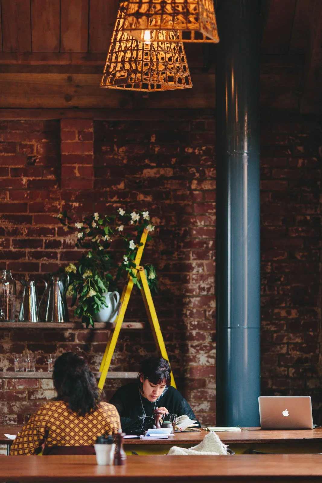 Two women sitting at a wooden table in a cozy café with exposed brick walls, one reading a book and the other looking at her phone. There are a laptop, books, a cup, and condiments on the table. A yellow ladder with flowers is in the background, and hanging basket-style lamps provide warm lighting.