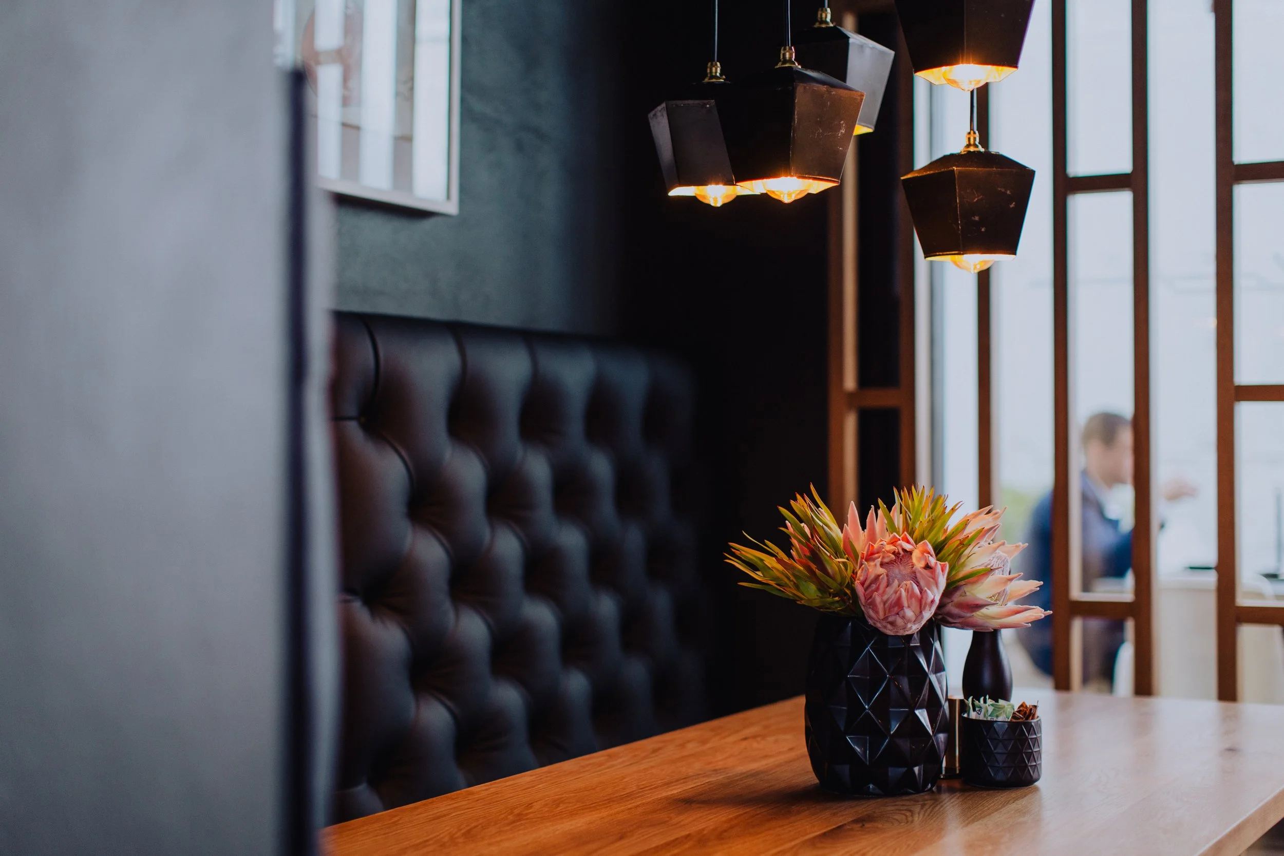 A cozy restaurant interior with pendant lighting, a wooden table, a black tufted leather booth, a vase with pink flowers, and a blurred person sitting by large windows.
