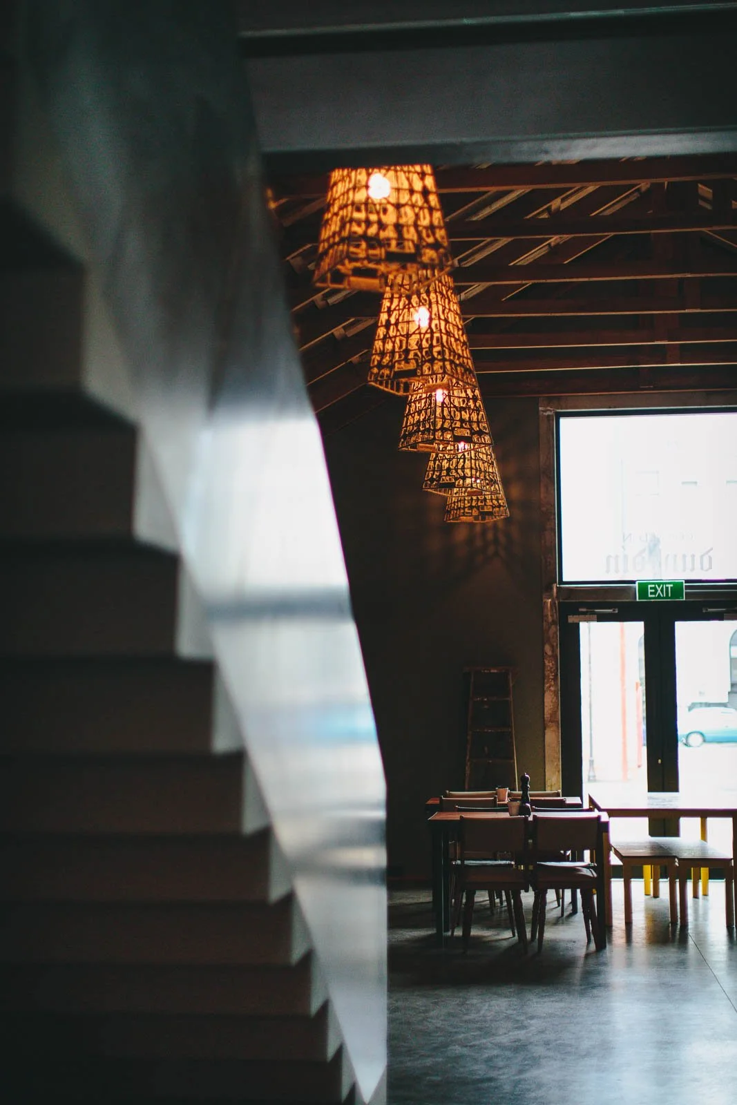 Interior of a modern restaurant or cafe with hanging pendant lights, a staircase on the left, tables, and chairs near the entrance, with a large window and an illuminated exit sign.
