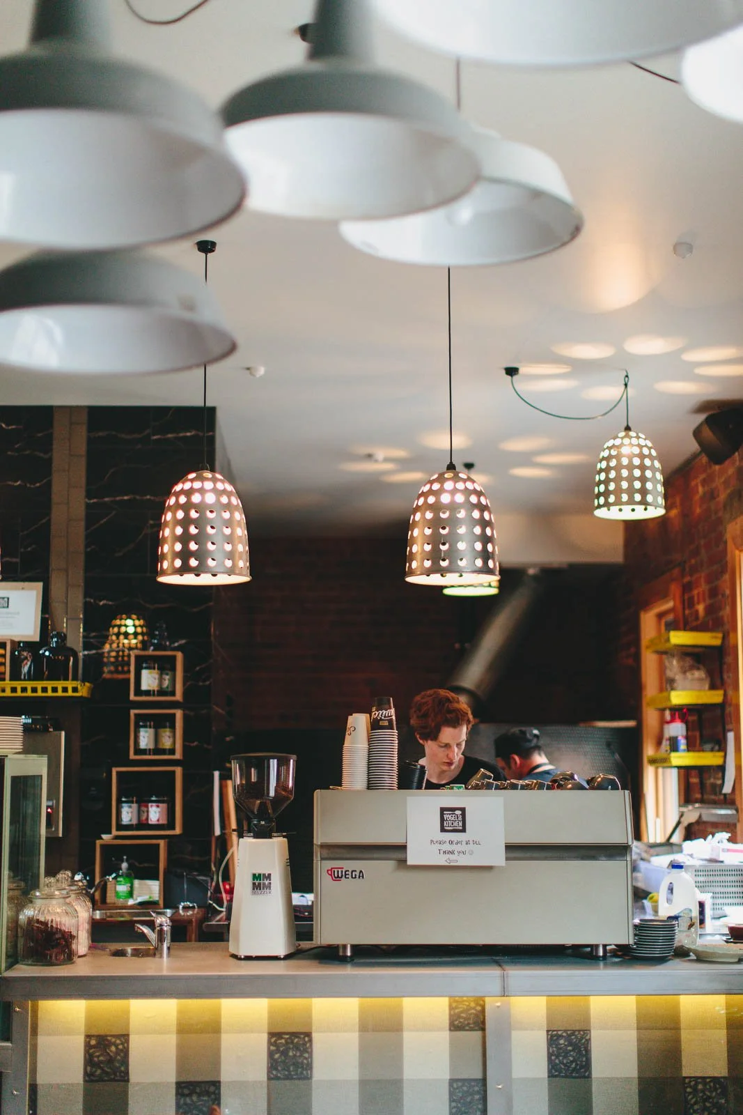 Interior of a coffee shop with hanging light fixtures, a barista working behind the counter, and shelves with cups and supplies.